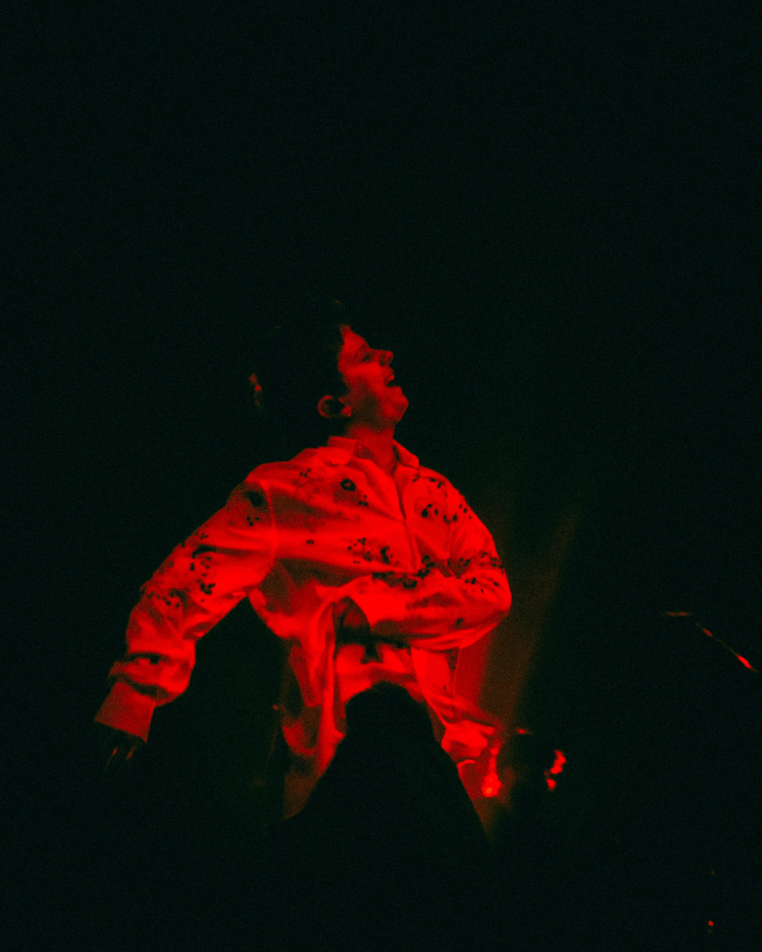 Performer on stage illuminated by red lights, wearing an embroidered shirt, with arms crossed, looking upward in a dark environment.