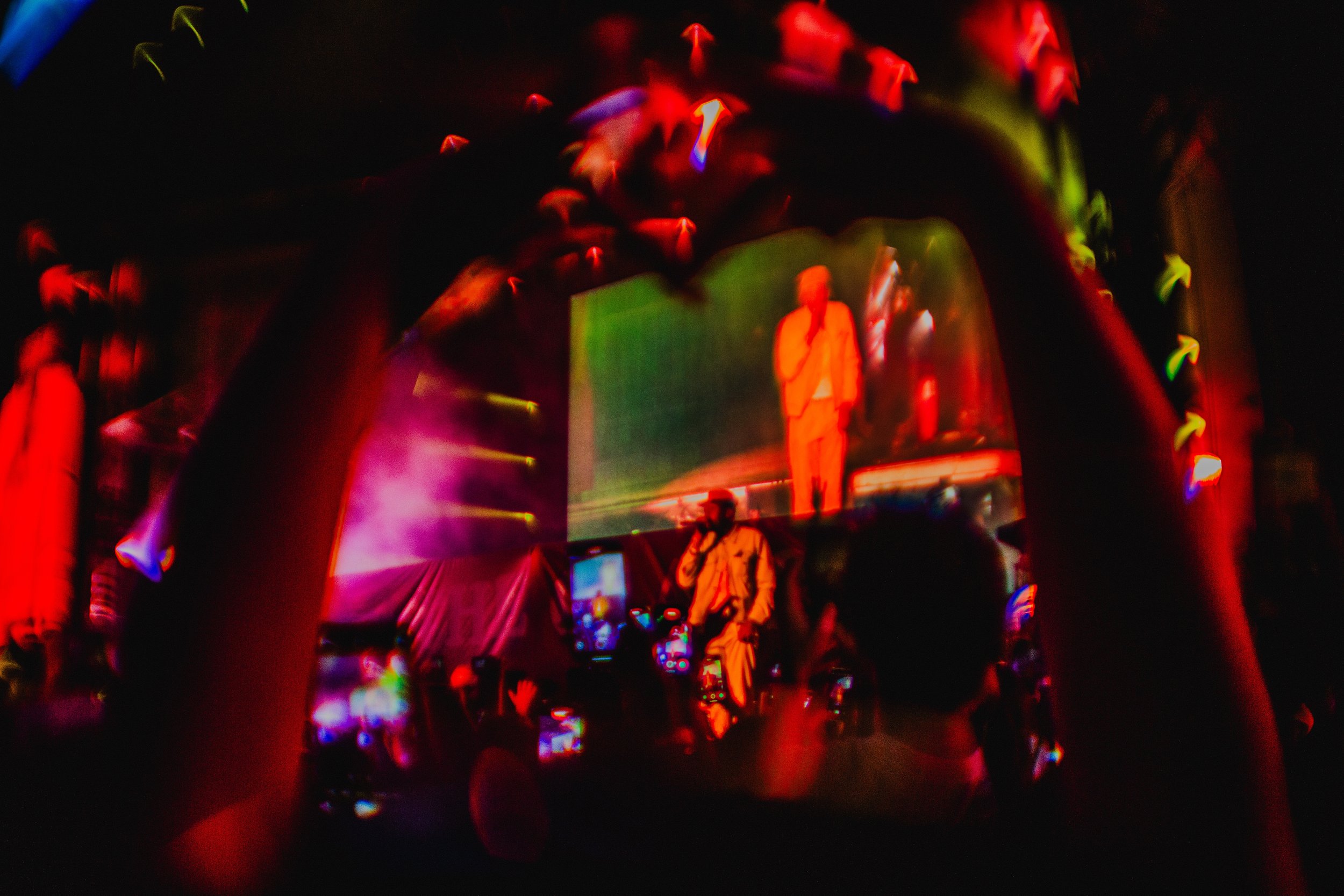 Person holding smartphone capturing a live performance on stage with colorful lighting, reflected on the phone screen showing a performer in a white suit, with a large screen projecting the same performer behind.