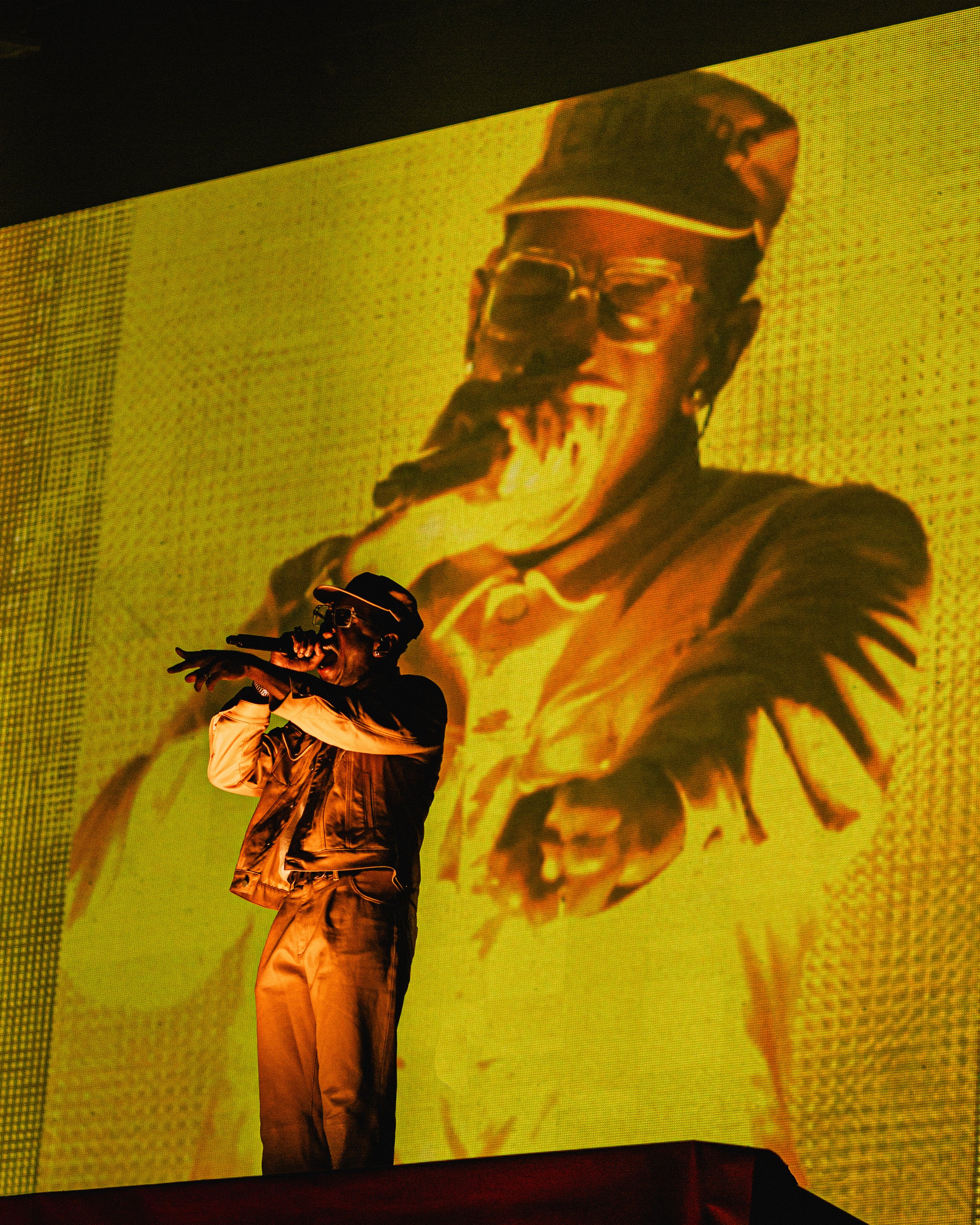 A performer on stage holding a microphone, with a large yellow portrait of a person singing projected behind them.
