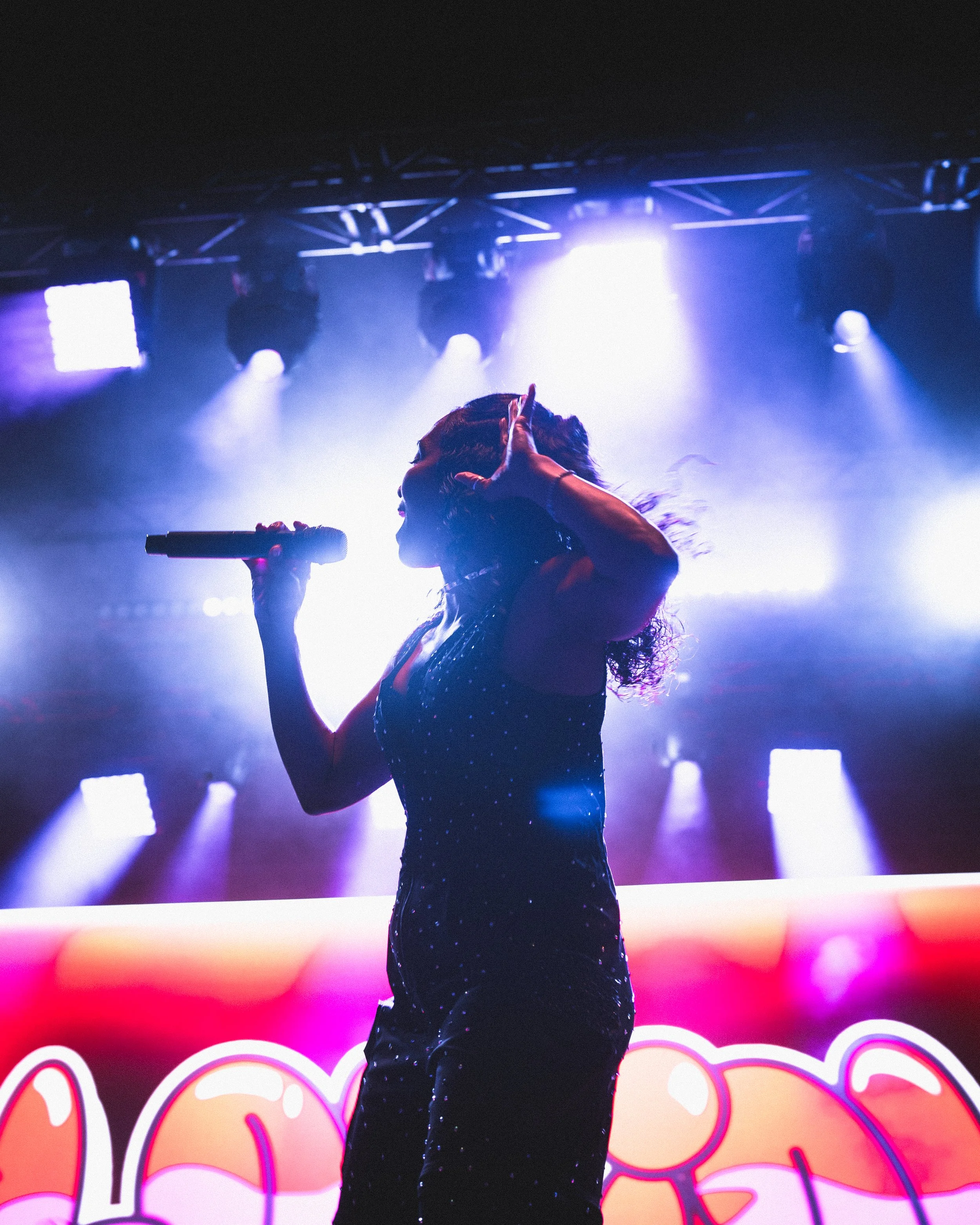 A singer with curly hair wearing a black dress with white dots, holding a microphone in one hand and making a hand gesture on her head, performing on stage with colorful lights and graffiti background.