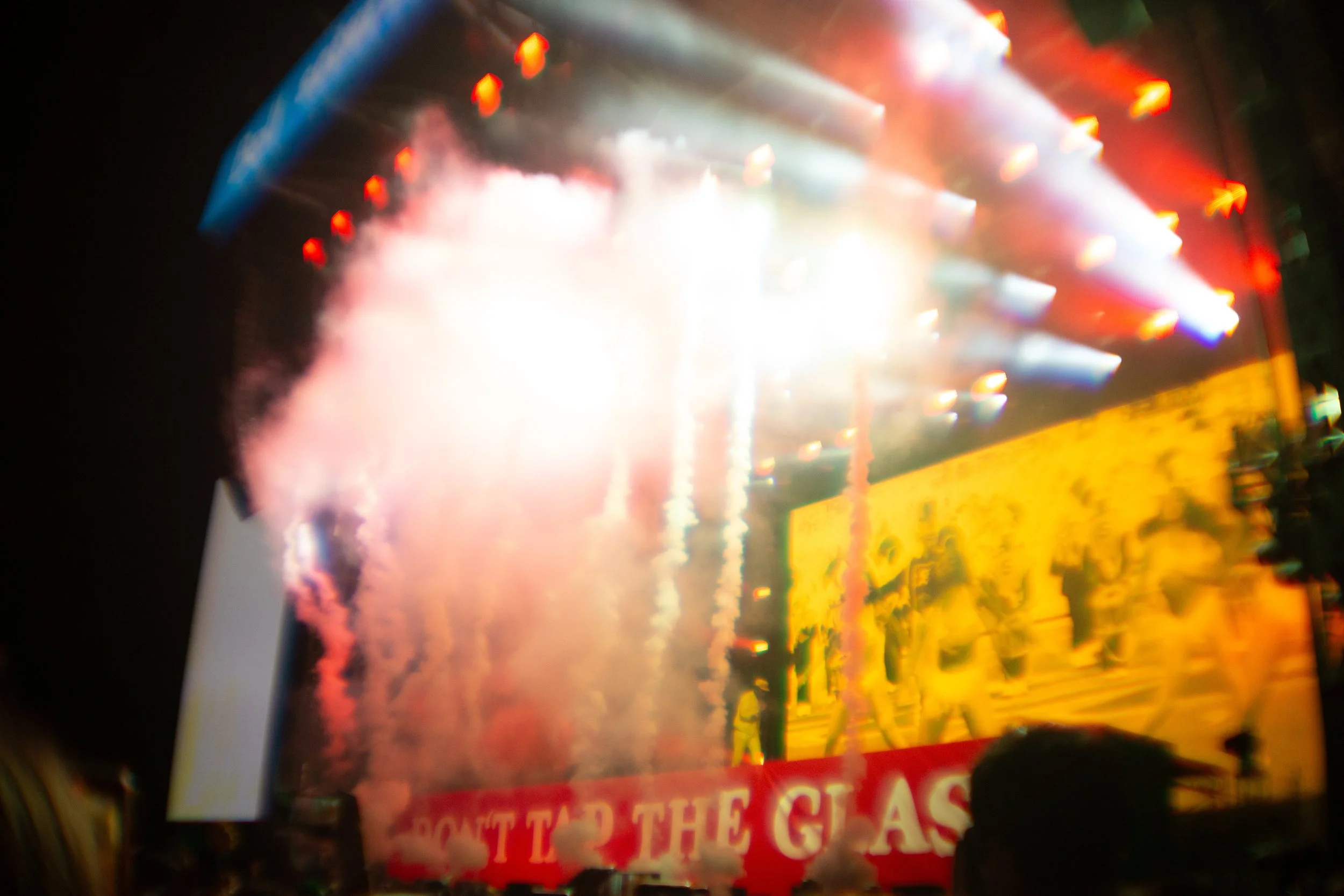 Bright fireworks display with smoke and colorful lights at night, with a partially visible yellow and red banner reading 'DON'T TAP THE GLASS'.
