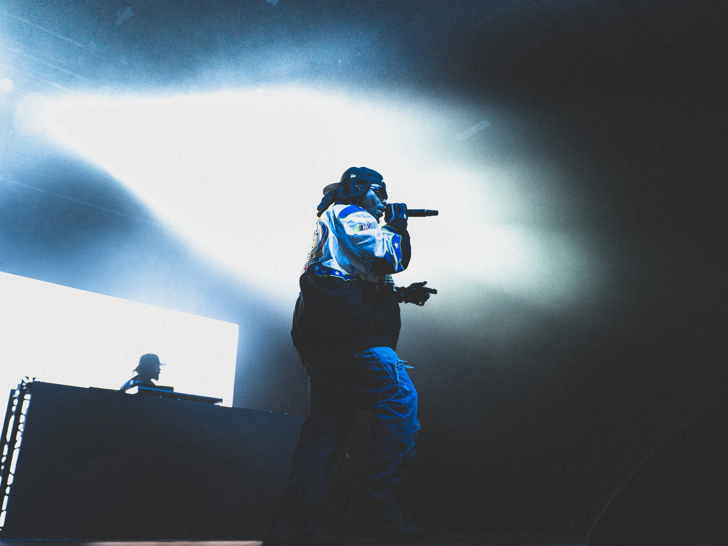 A performer on stage holding a microphone, wearing sunglasses, a cap, and a blue and black jacket, with a DJ in the background.