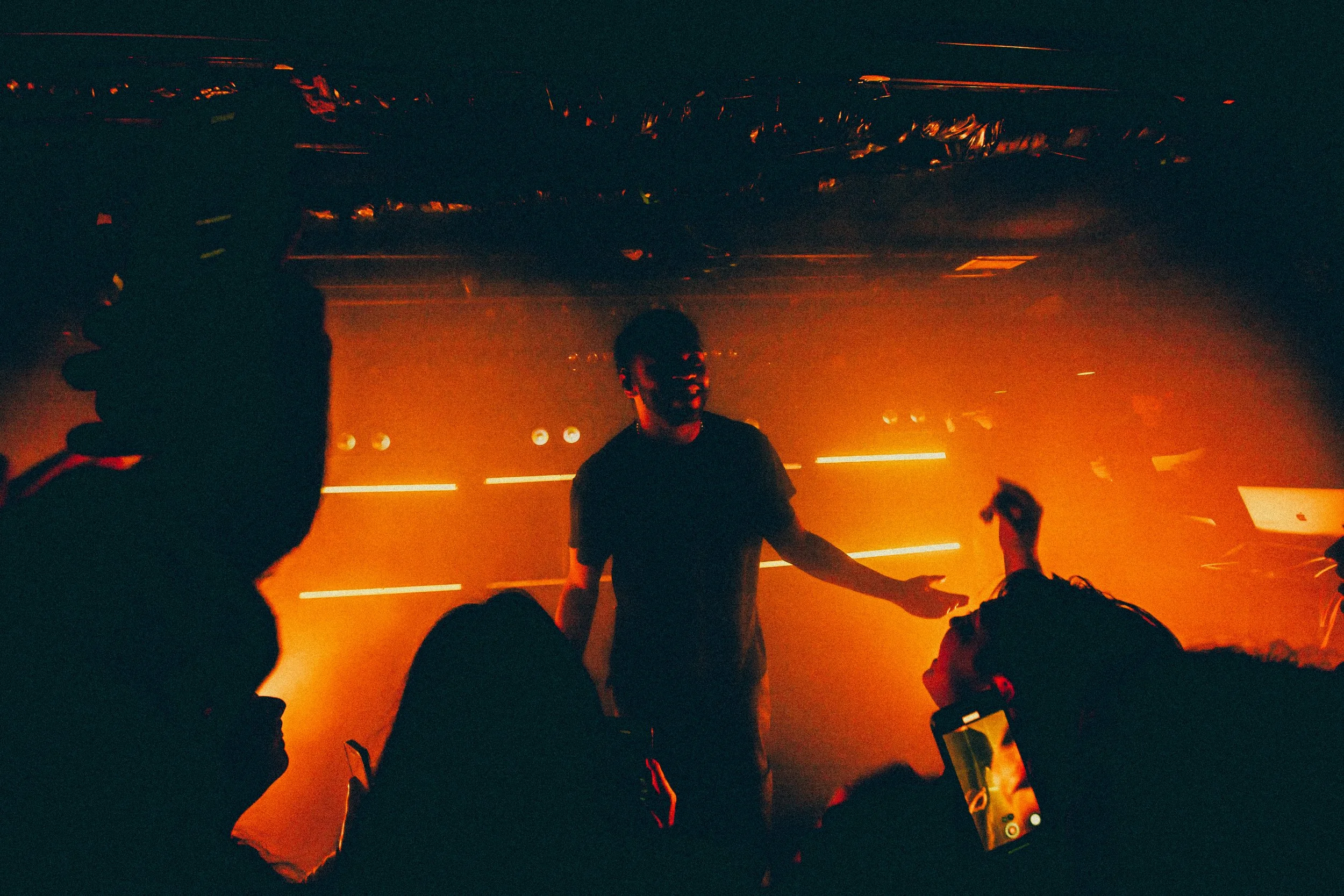 A DJ performing at a concert with orange stage lighting, silhouetted crowd in the foreground, and someone taking a photo with a smartphone.