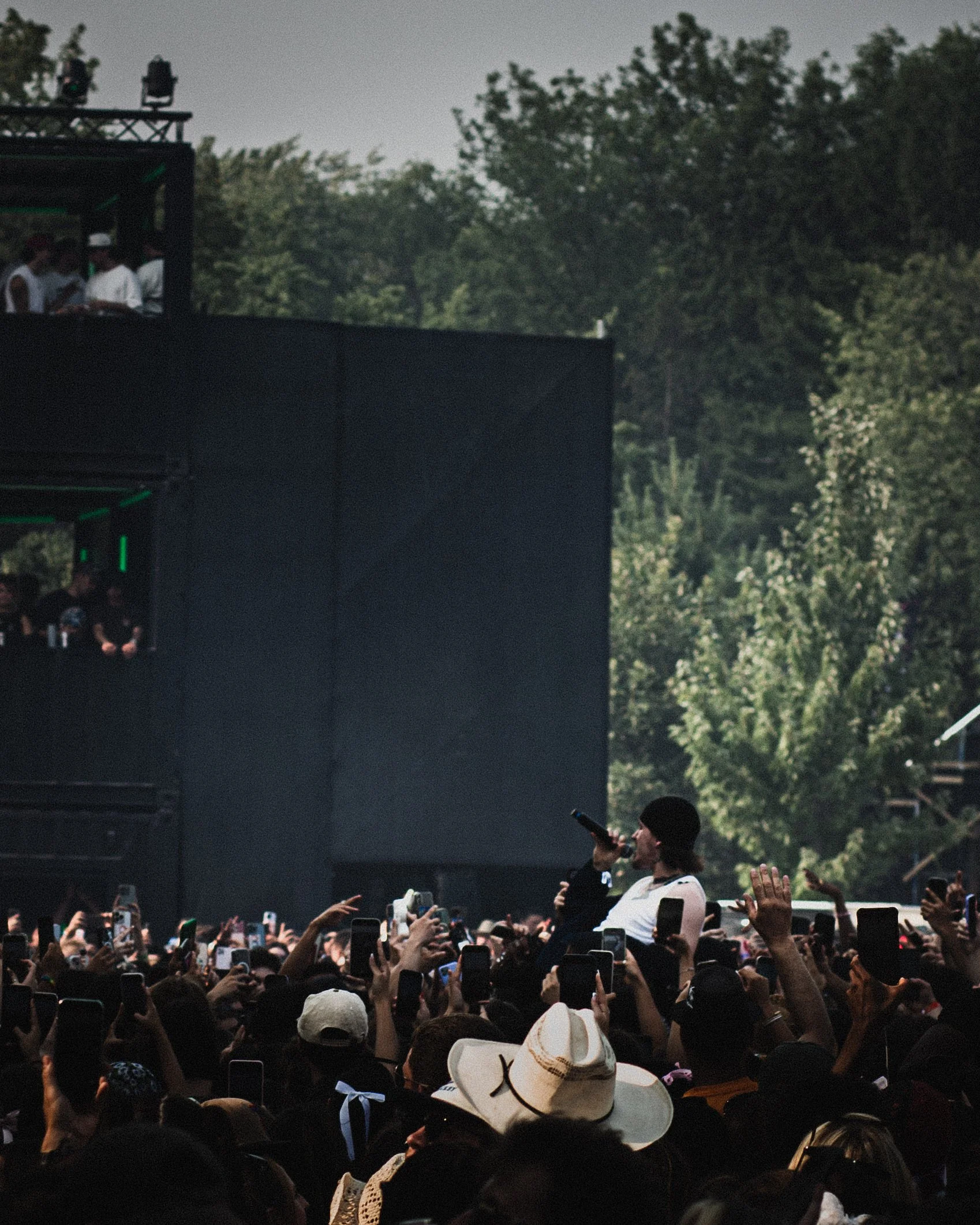 Performer singing on stage at an outdoor concert with a large crowd taking pictures and videos.