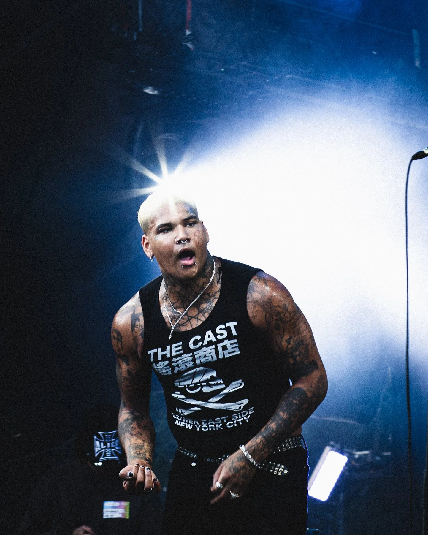 A tattooed person with short blonde hair singing or performing on stage, wearing a black shirt that says 'The Cast' and featuring a graphic of a burger, with bright stage lights behind them.