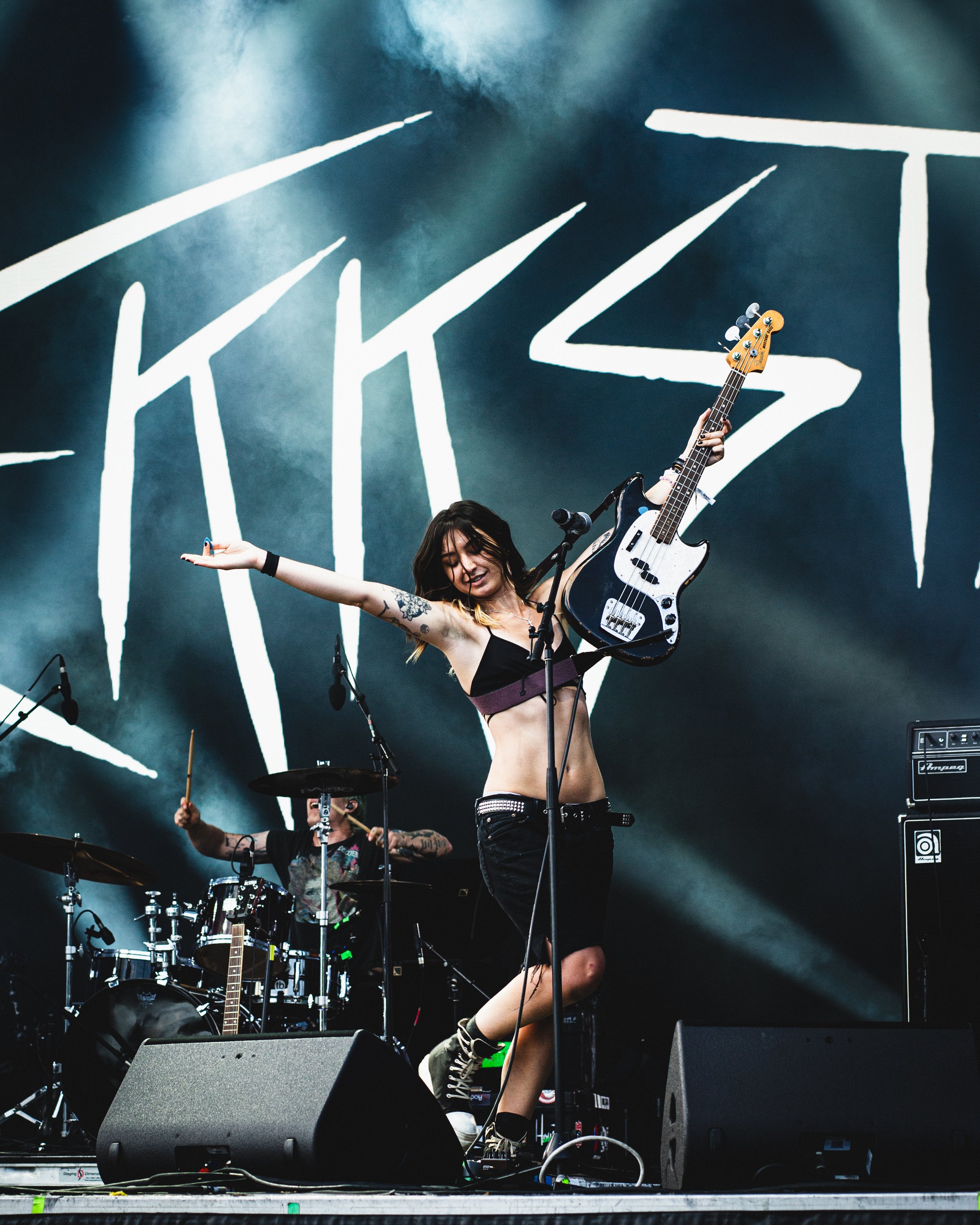 A female musician holding a bass guitar on stage during a live concert, with a drummer playing in the background and a large stylized logo projected behind them.