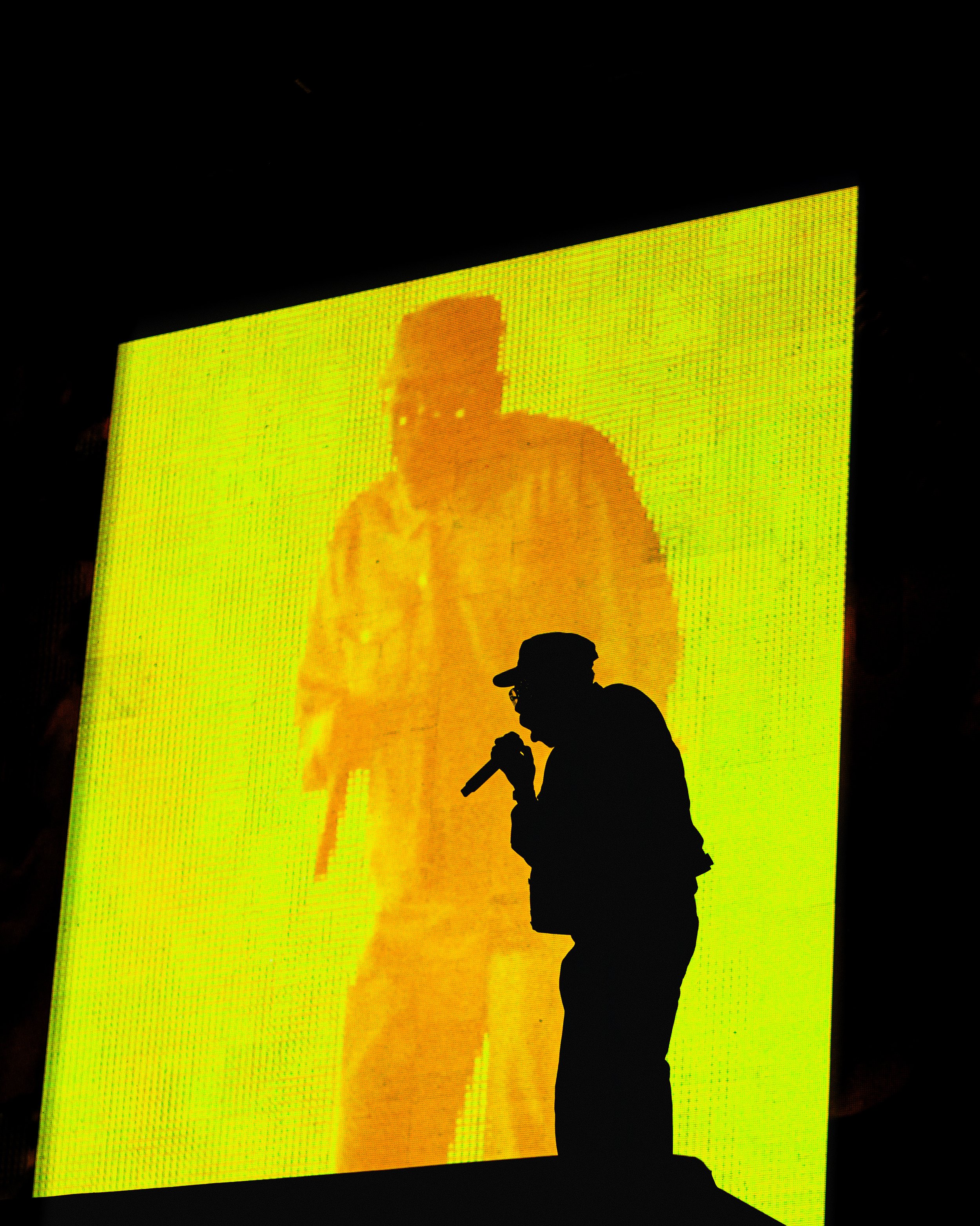 Silhouette of a man holding a microphone on stage in front of a large yellow screen displaying a pixelated image of another person.