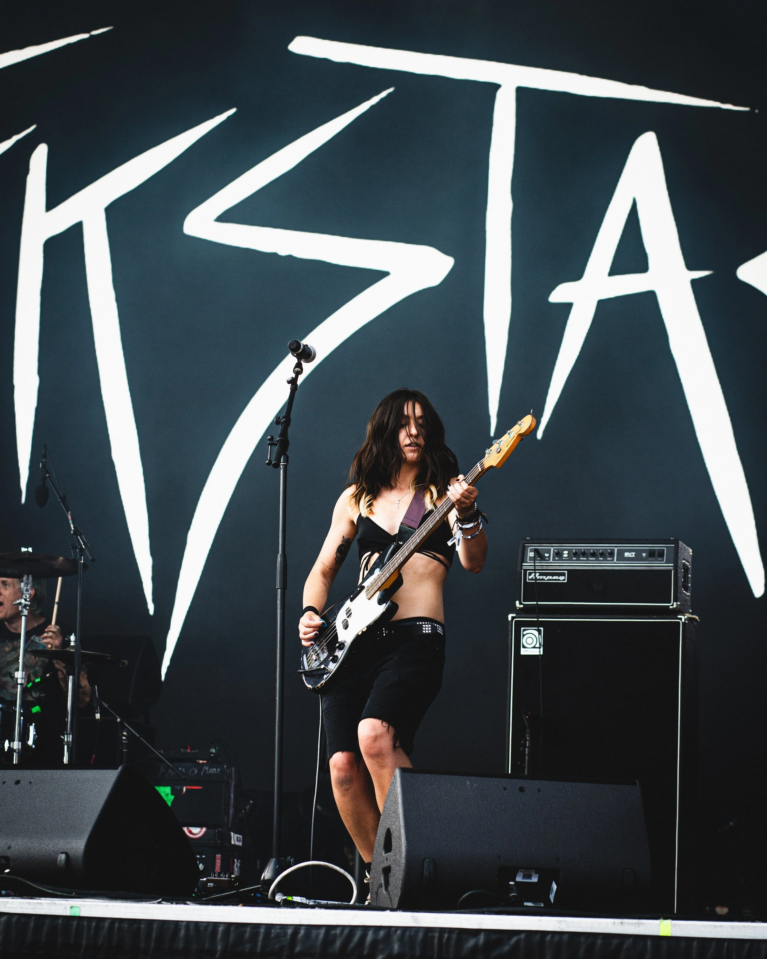 Female musician playing electric guitar on stage during a live concert with a large stylized logo behind her.
