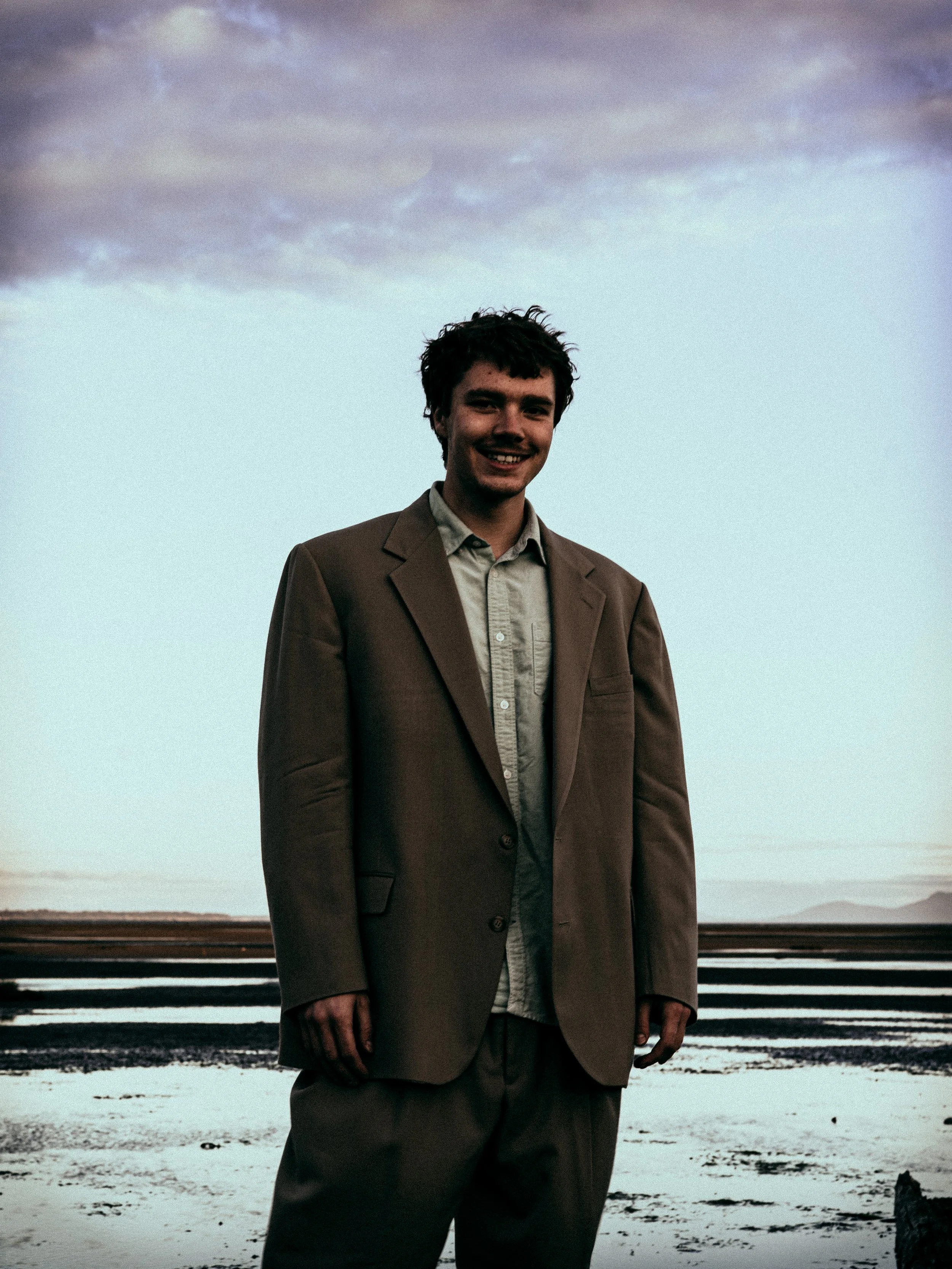 A young man standing outdoors by the water during sunset, wearing a brown blazer and beige pants, smiling at the camera.