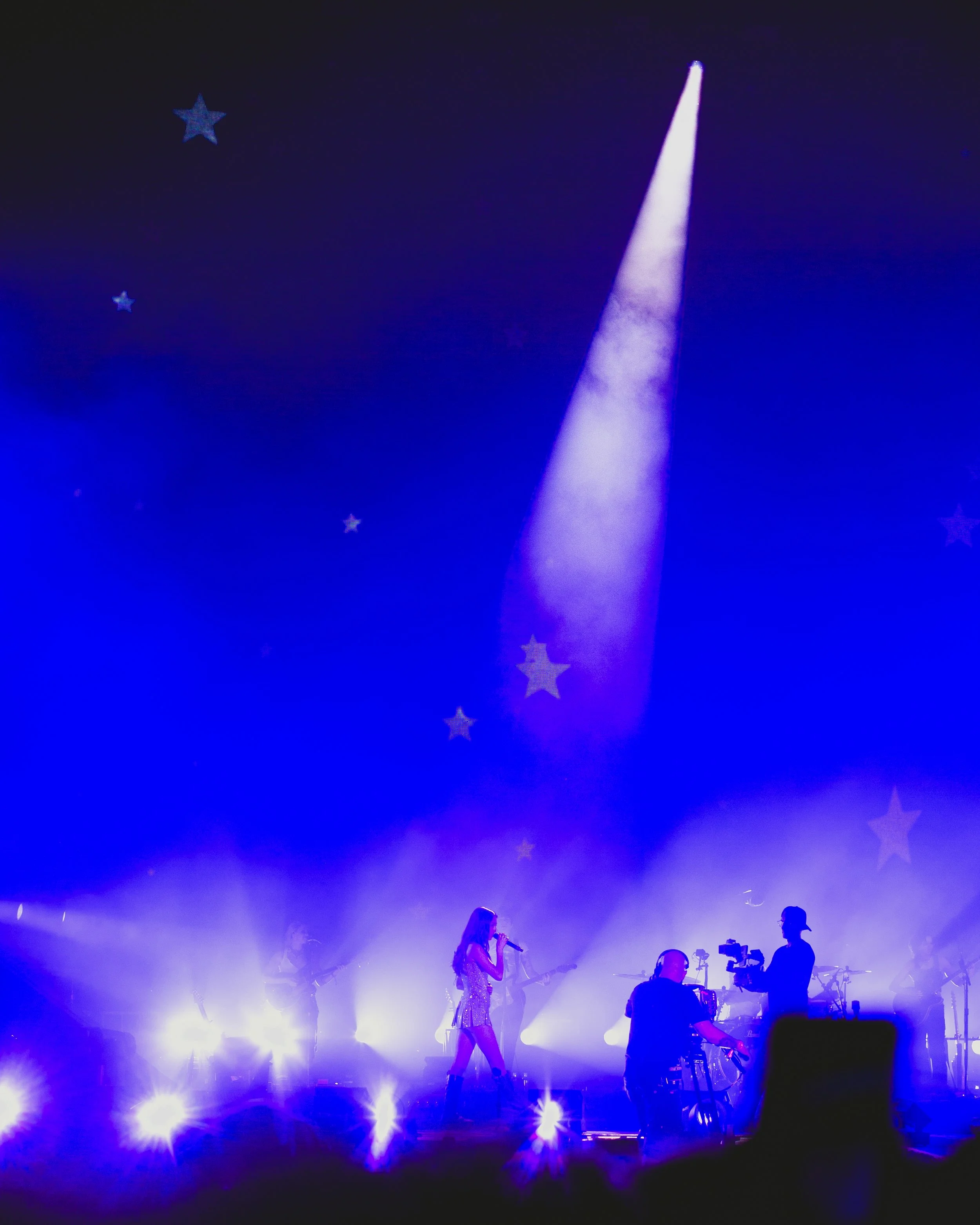A singer performs on stage with a band, illuminated by colorful stage lights and a blue backdrop with star decorations, with camera operators capturing the performance.