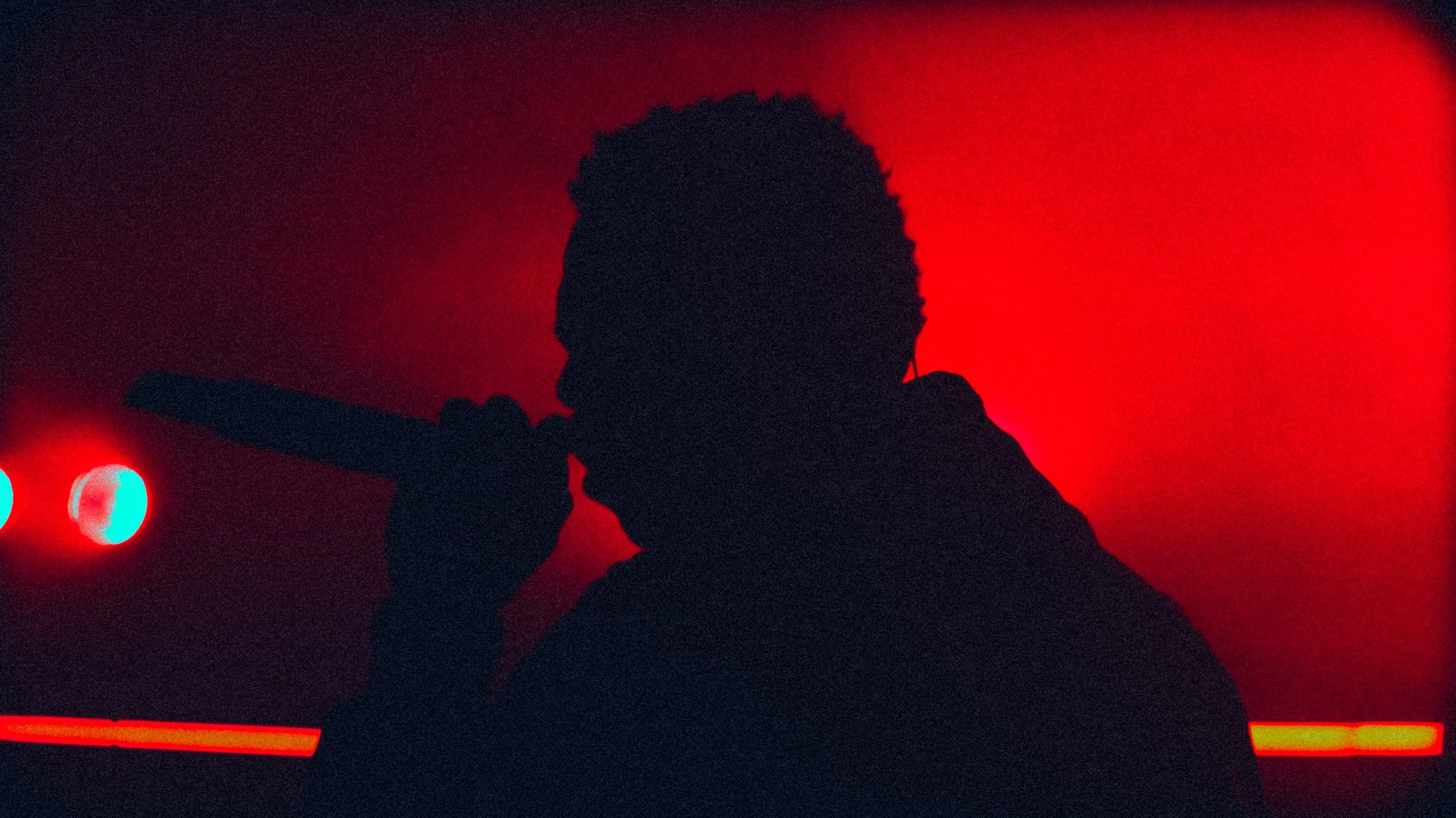 Silhouette of a person singing into a microphone with a red background and multicolored stage lights.