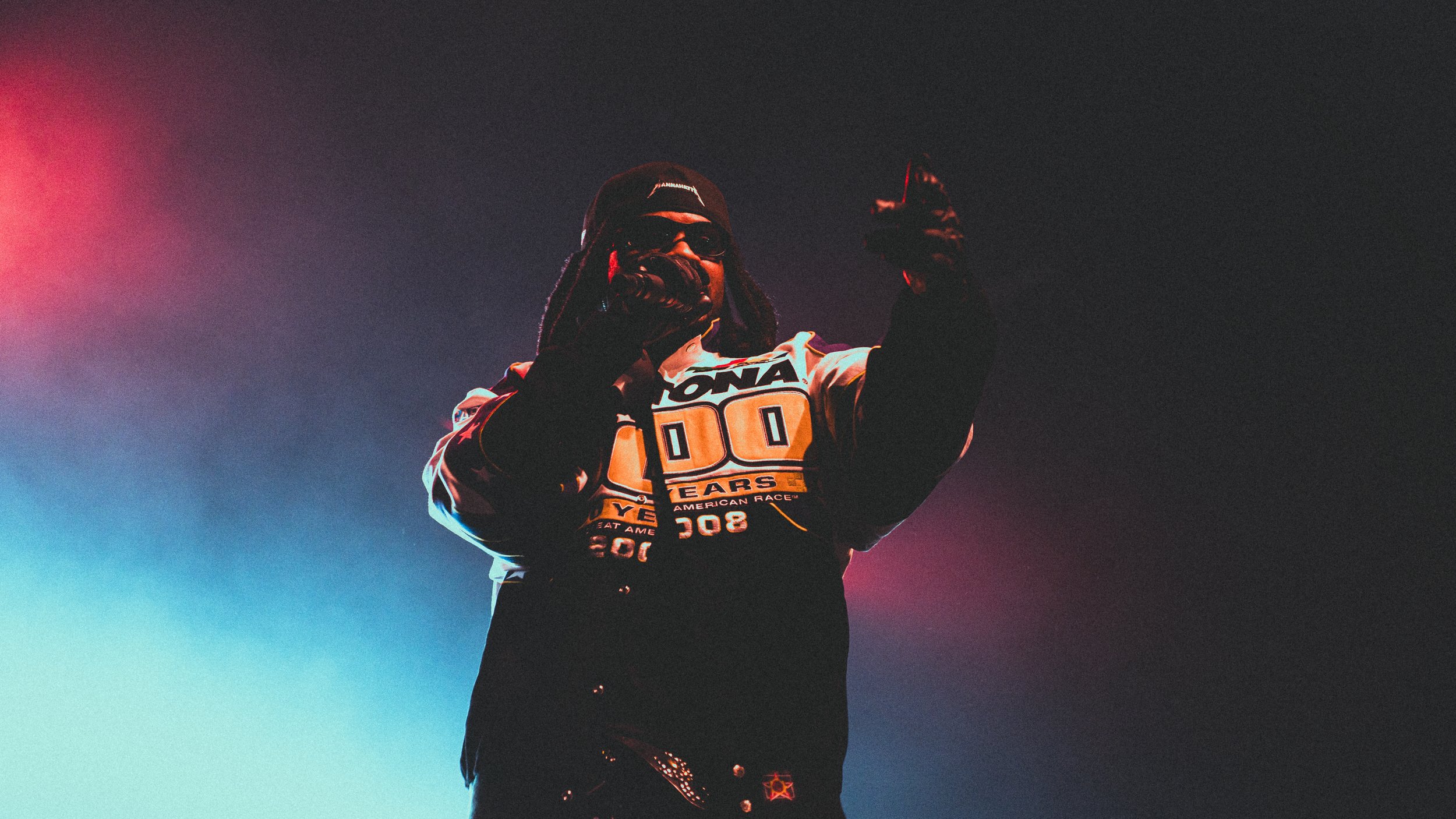 A person wearing sunglasses, a beanie, and gloves, dressed in a racing jacket with patches, giving a thumbs-up against a dark background with colorful lighting.