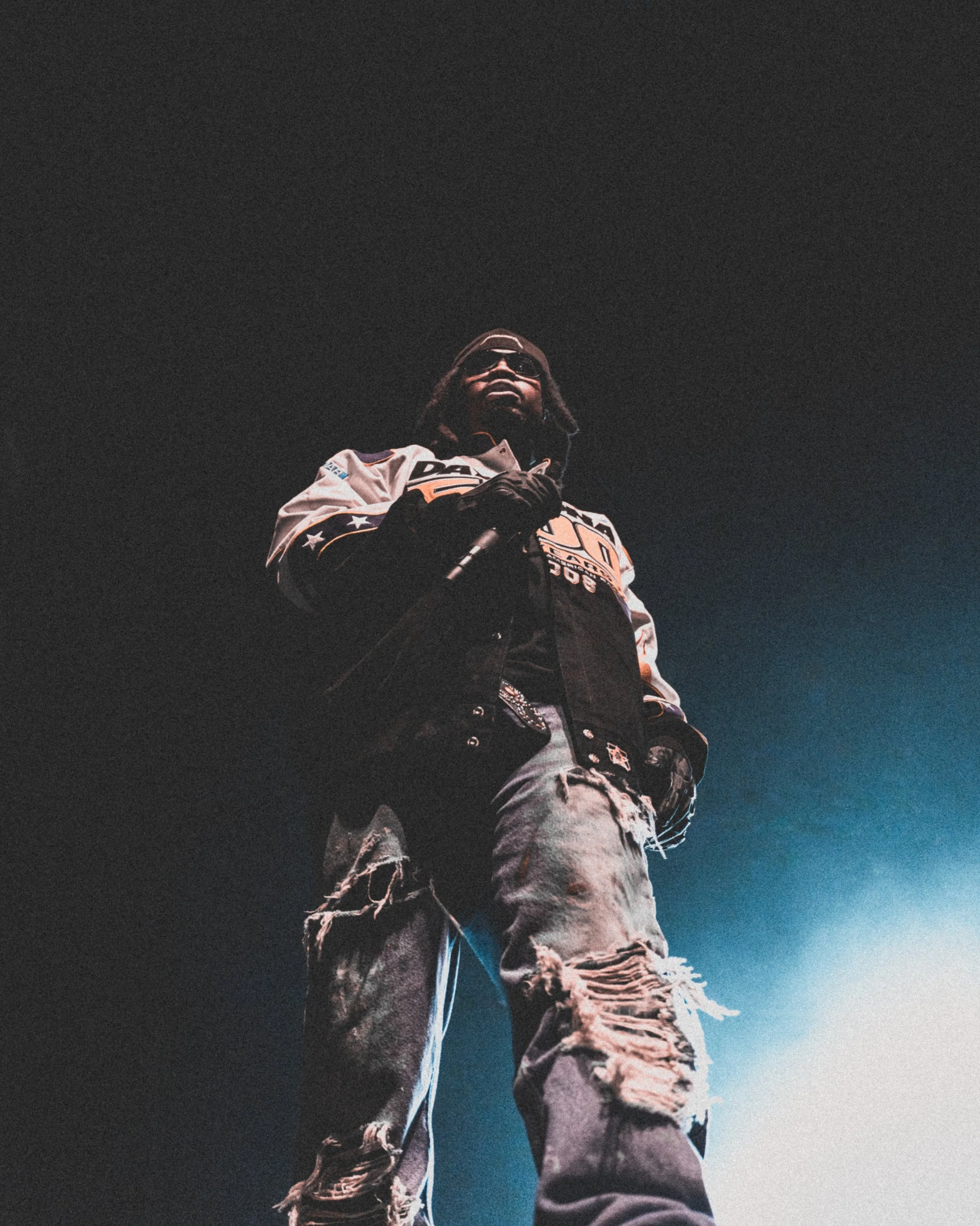 A person standing in a dark environment, wearing sunglasses, a racing-style jacket, and ripped jeans, holding a microphone.