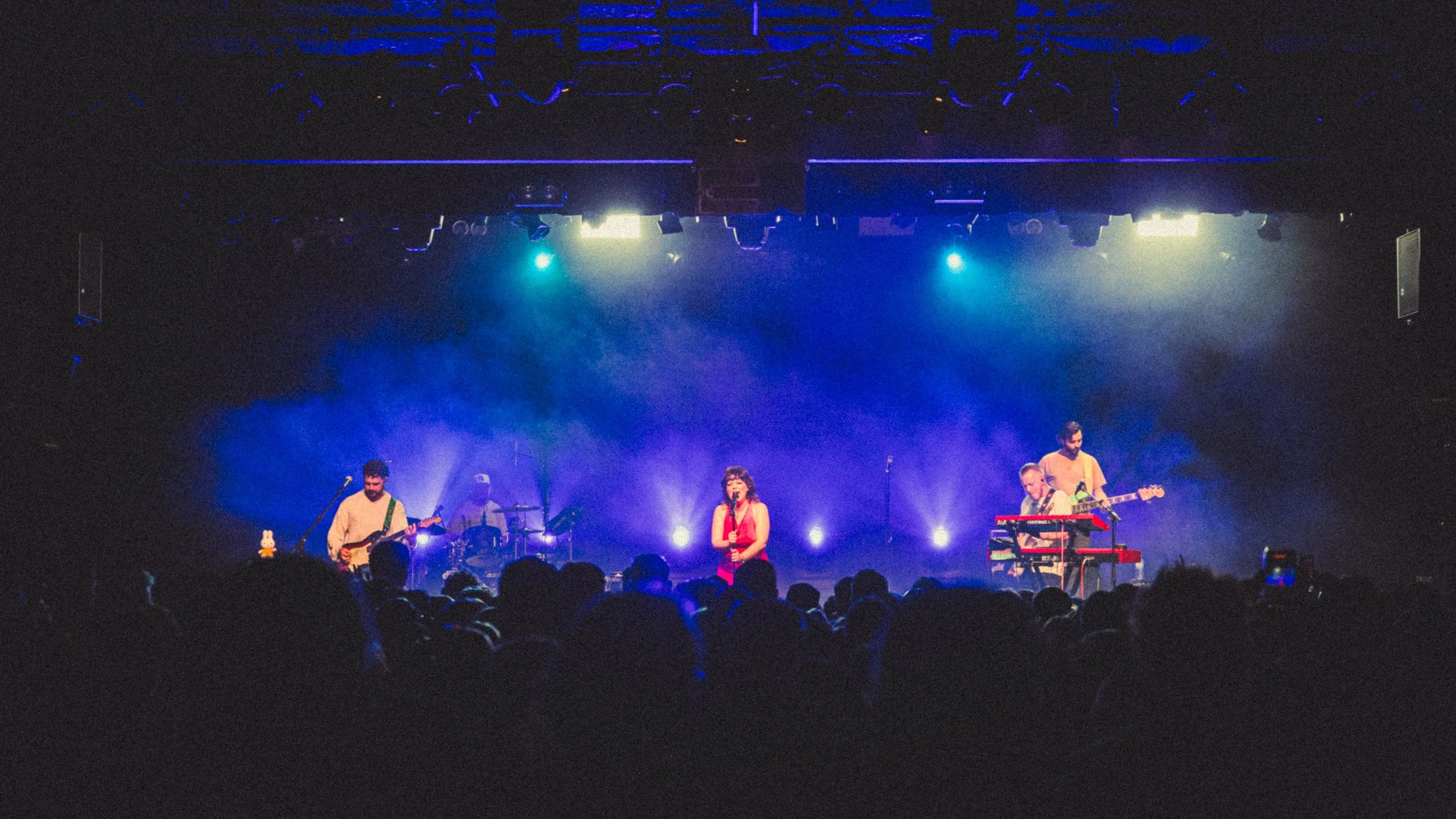 A live concert with a band playing on stage, including a female singer, drummer, and two guitarists, illuminated by colorful stage lights, with an audience in the foreground.