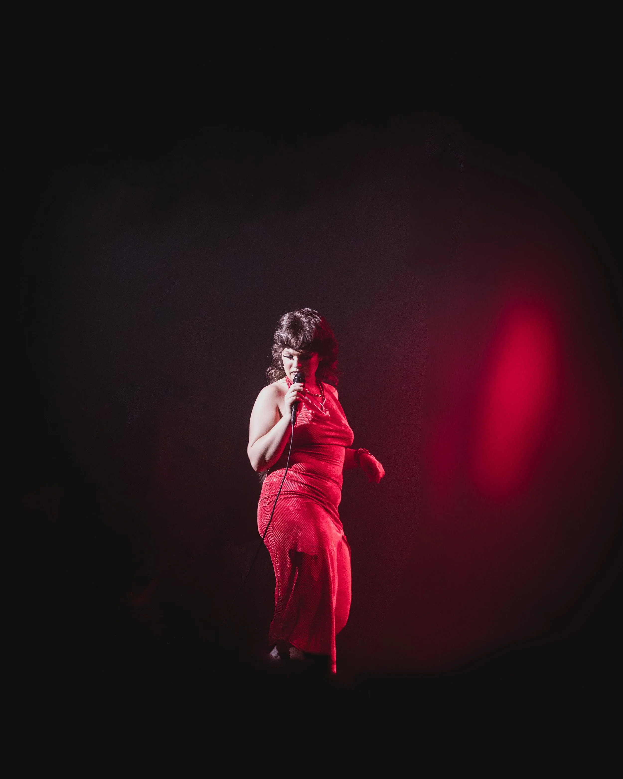 A woman in a red dress singing into a microphone on stage, illuminated by a pink spotlight against a dark background.