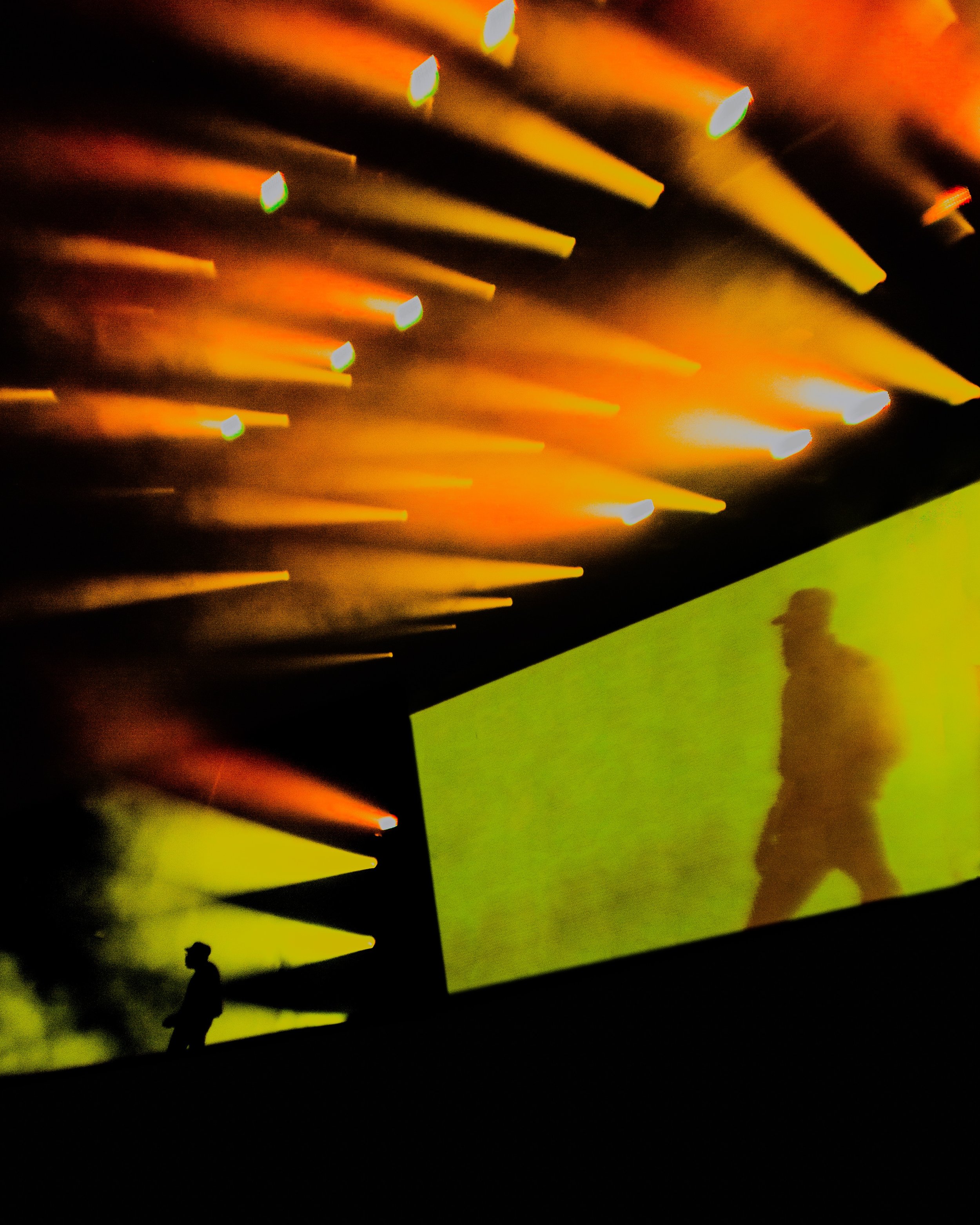 Stage with silhouetted person in front of large screen showing shadow of a walking figure, colorful stage lighting with yellow and orange beams.