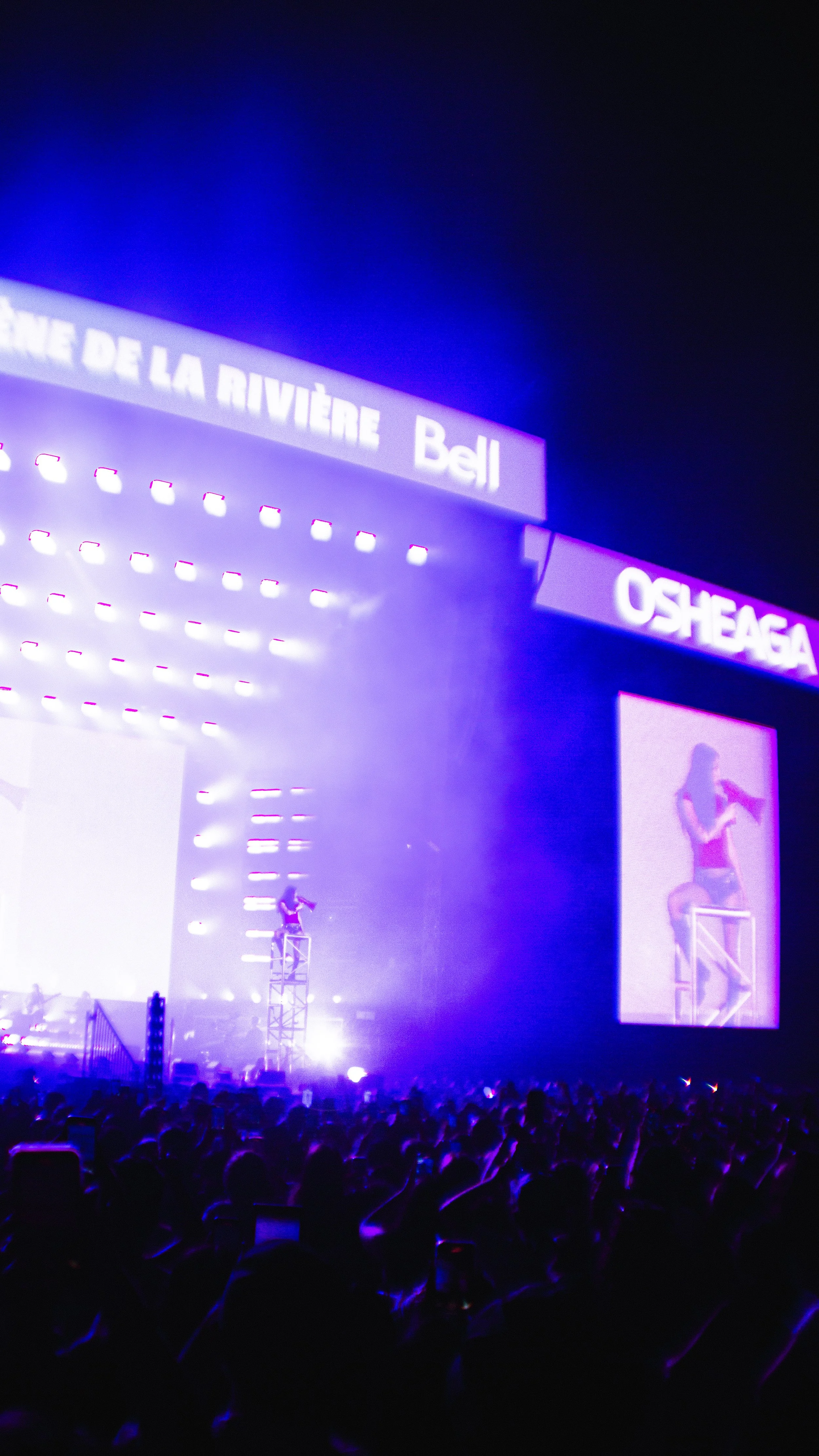 A concert stage illuminated with purple and blue lights, featuring a performer on a tall platform and large screens displaying the performance. Banners with the words "BIENVENUE DE LA RIVIÈRE Bell" and "OSHEAGA" are visible.