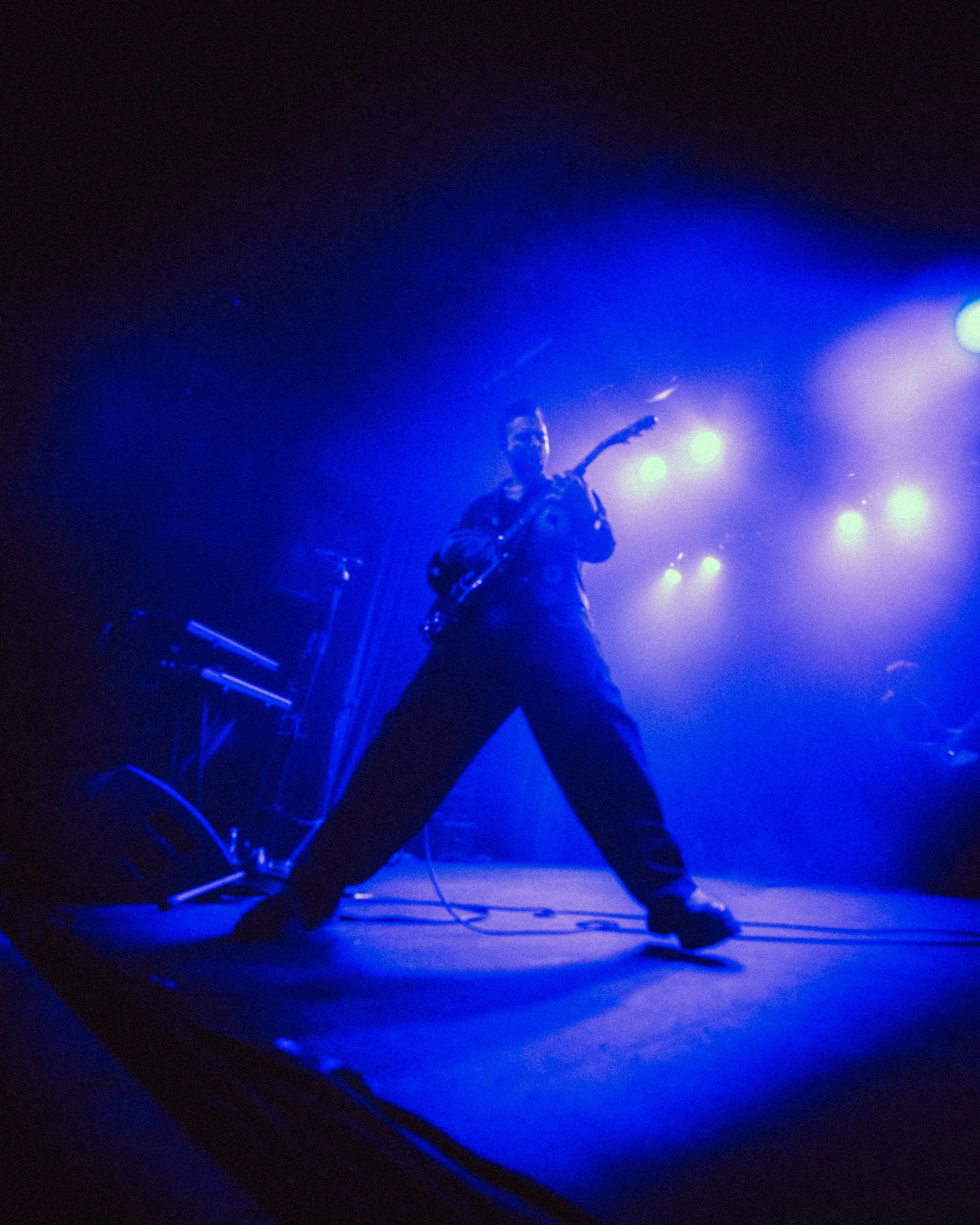 Musician playing a guitar on stage under blue lighting, with musical equipment visible in the background.