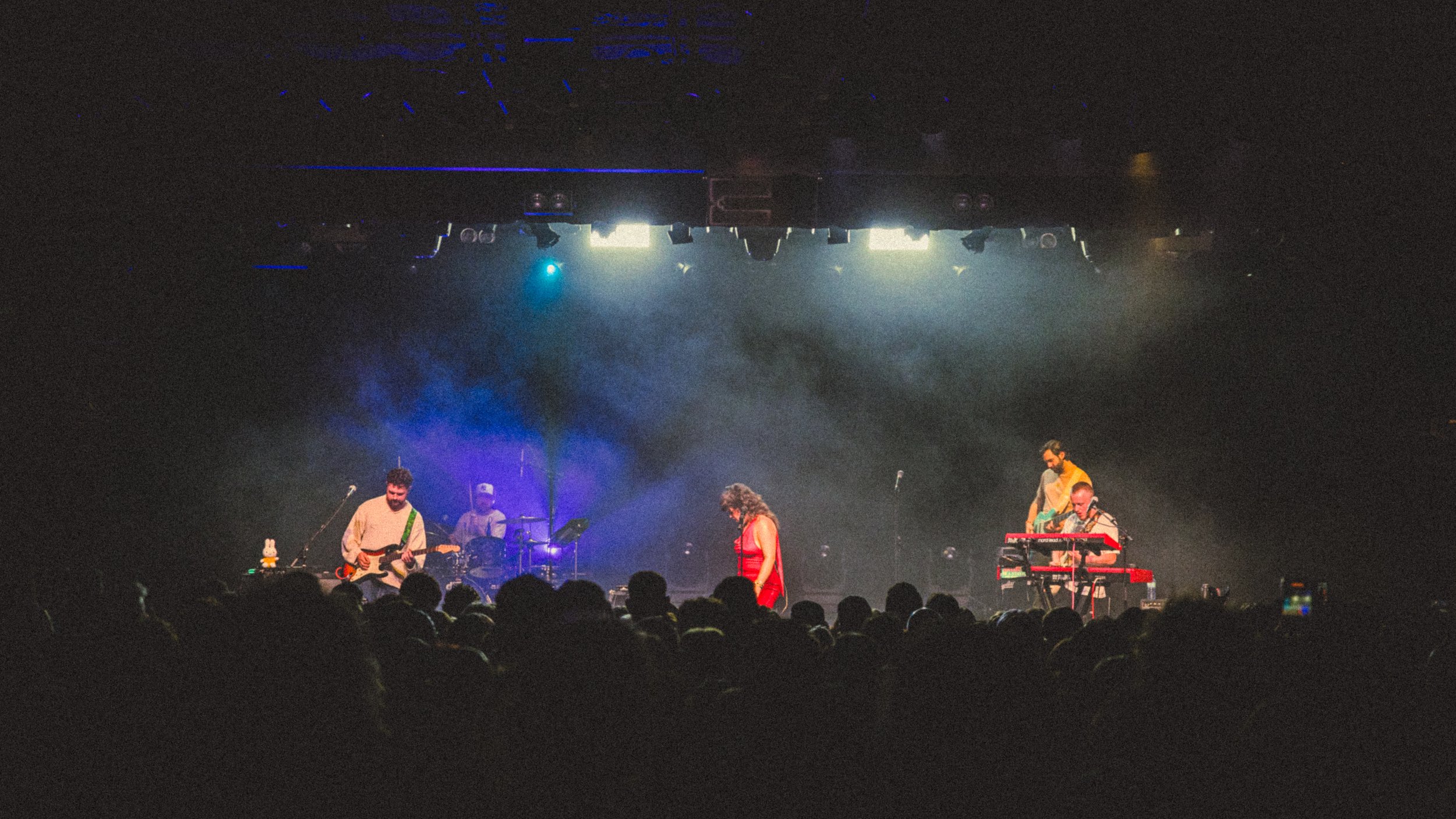 A band performing on stage with a vocalist, guitarist, drummer, and keyboard player, illuminated by stage lights, in front of a large audience.