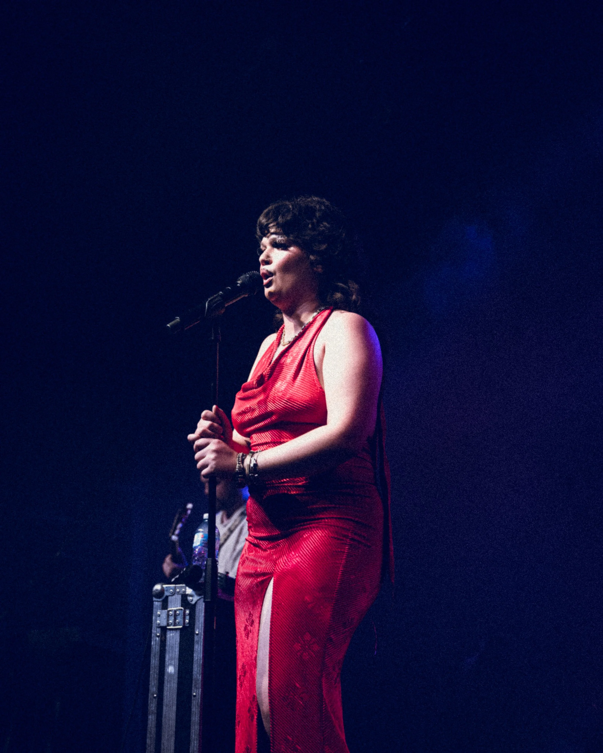 Performer singing into microphone on stage with dark background, wearing a red dress with high slit.
