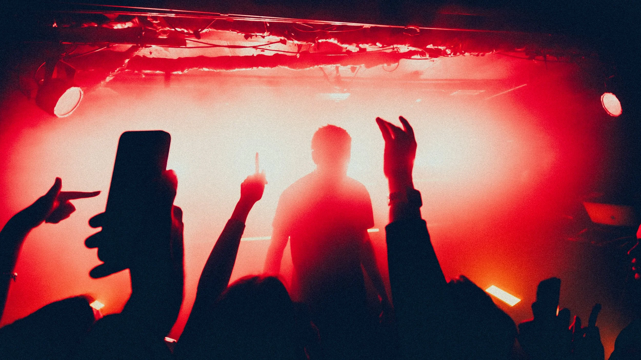 A concert scene with a performer on stage silhouetted against red lighting, with audience members raising their hands and taking photos.