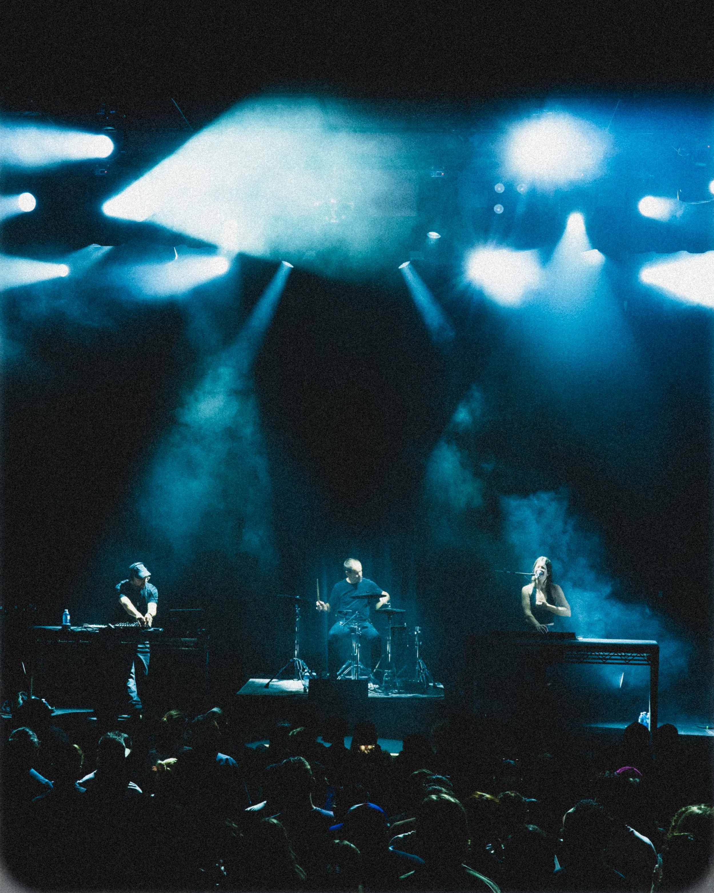 A live concert scene with a band performing on stage, illuminated by blue and white stage lights, with an audience watching in the foreground.