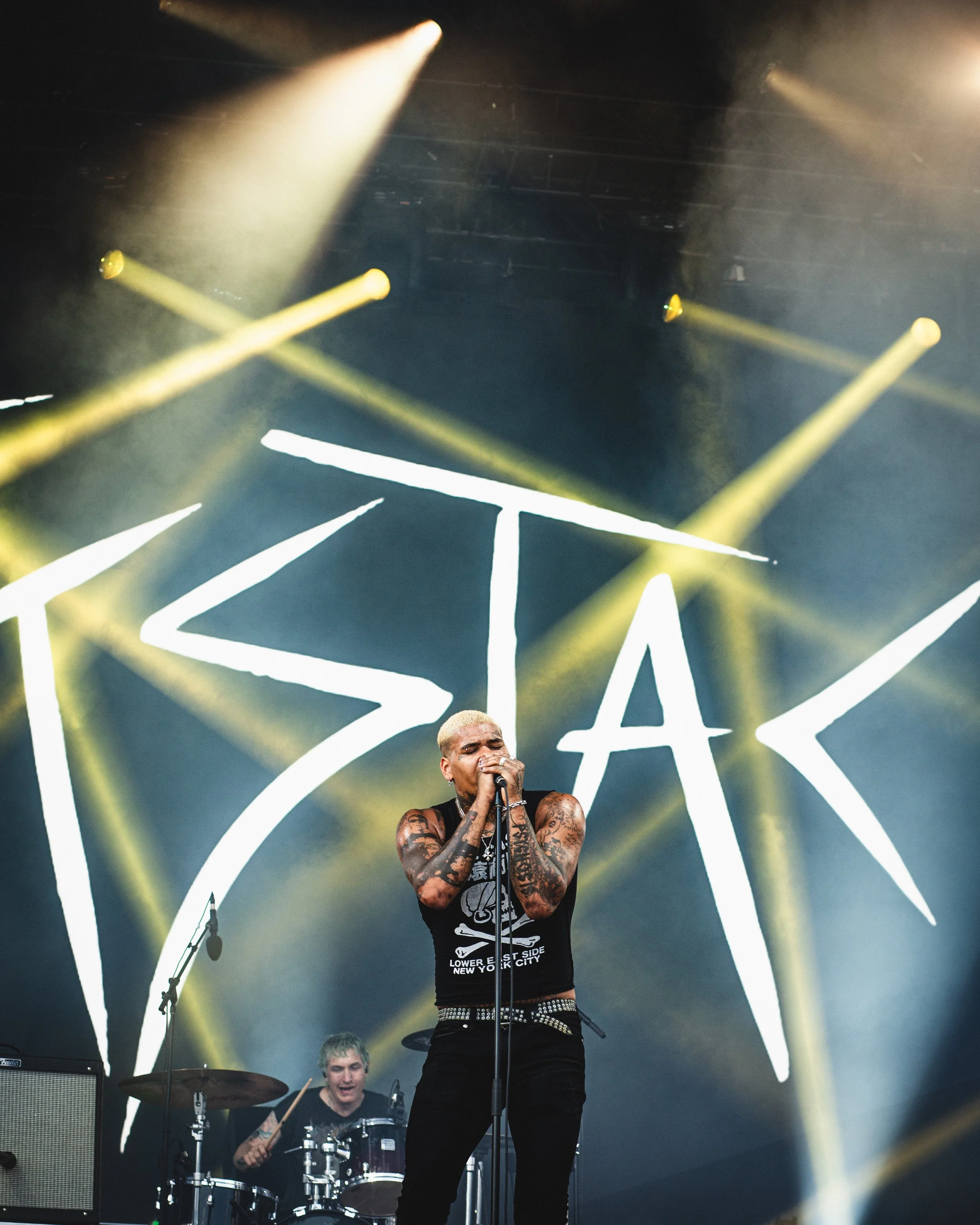 A musician with platinum blonde hair and tattoos performing on stage with a microphone, with a drummer in the background and a large "IZAK" sign projected behind.