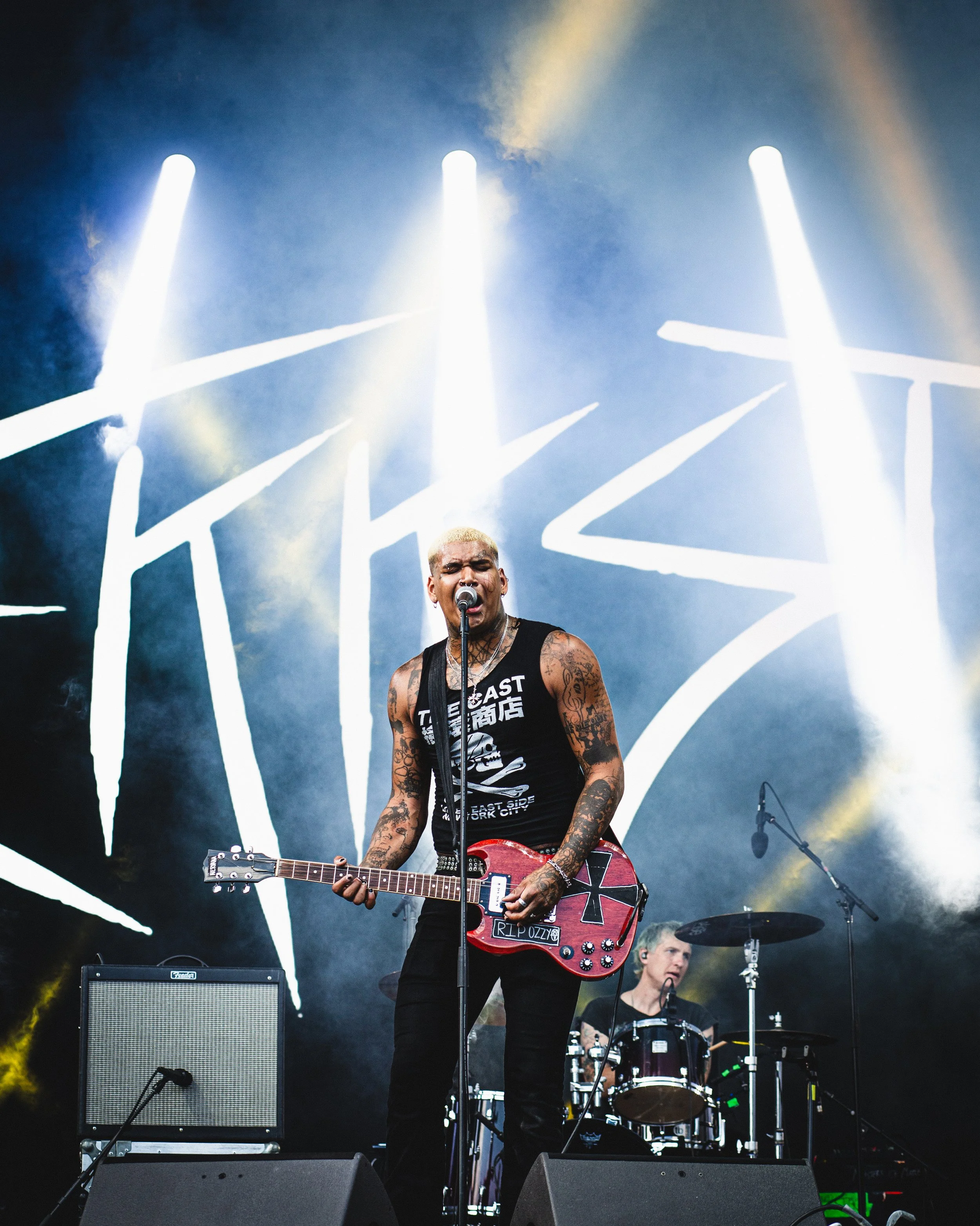 A vocalist with blonde hair, tattoos, and a sleeveless black shirt performs on stage with a red guitar, singing into a microphone. A drummer is seen in the background, playing drums, with a large black and white graphic behind them.