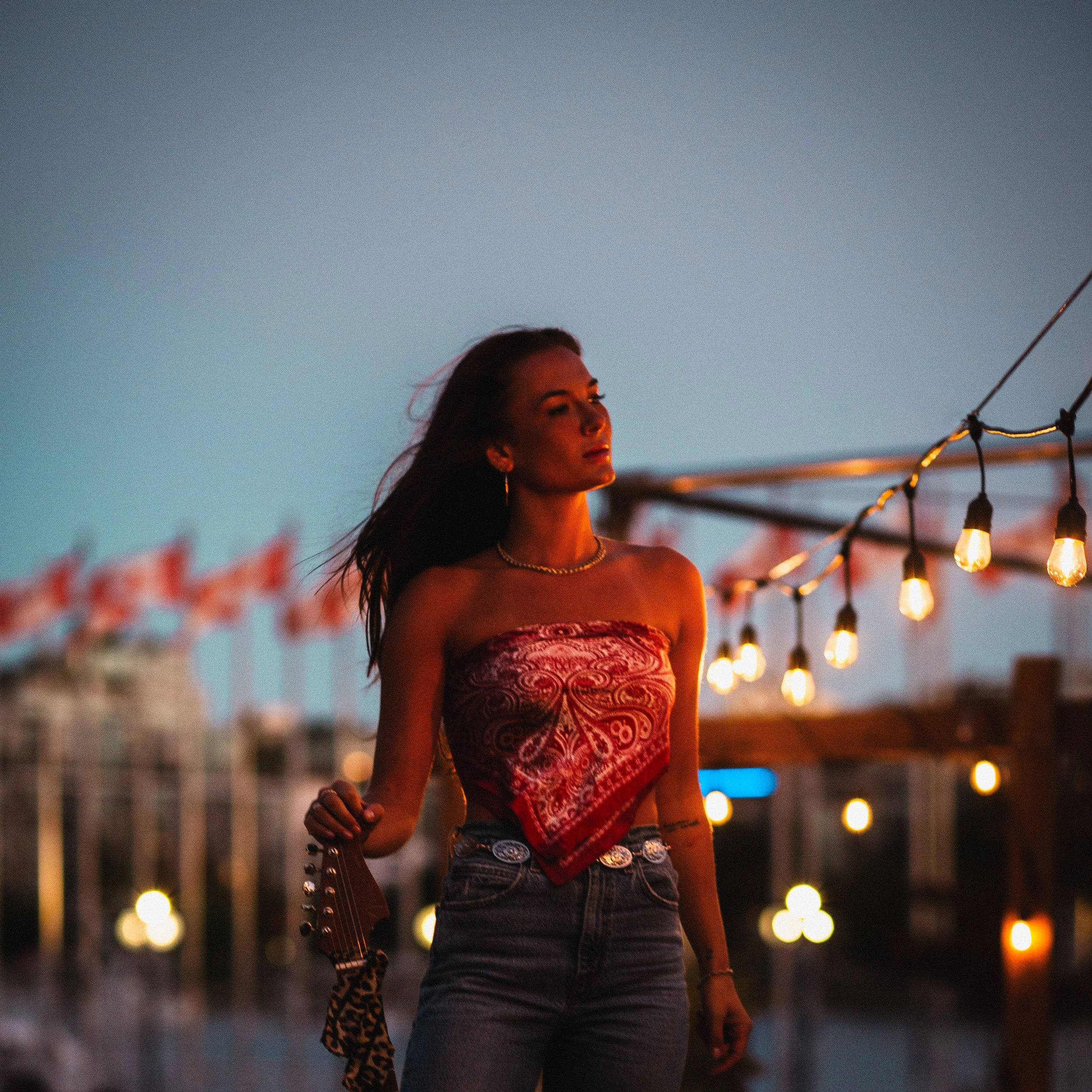 A young woman with long hair stands outdoors at dusk, wearing a red strapless top, jeans, and jewelry, next to string lights and a body of water in the background.