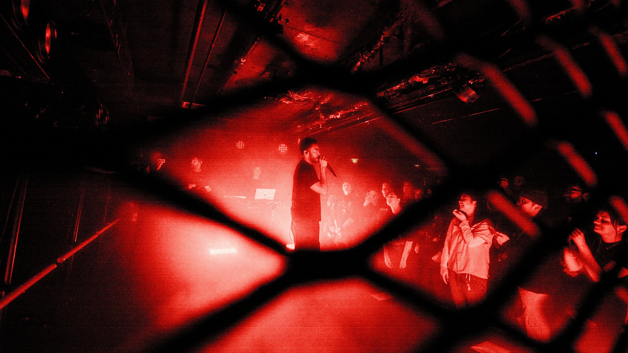 A performer singing into a microphone on stage at a concert, viewed through a chain-link fence with red lighting, with an audience watching in the background.