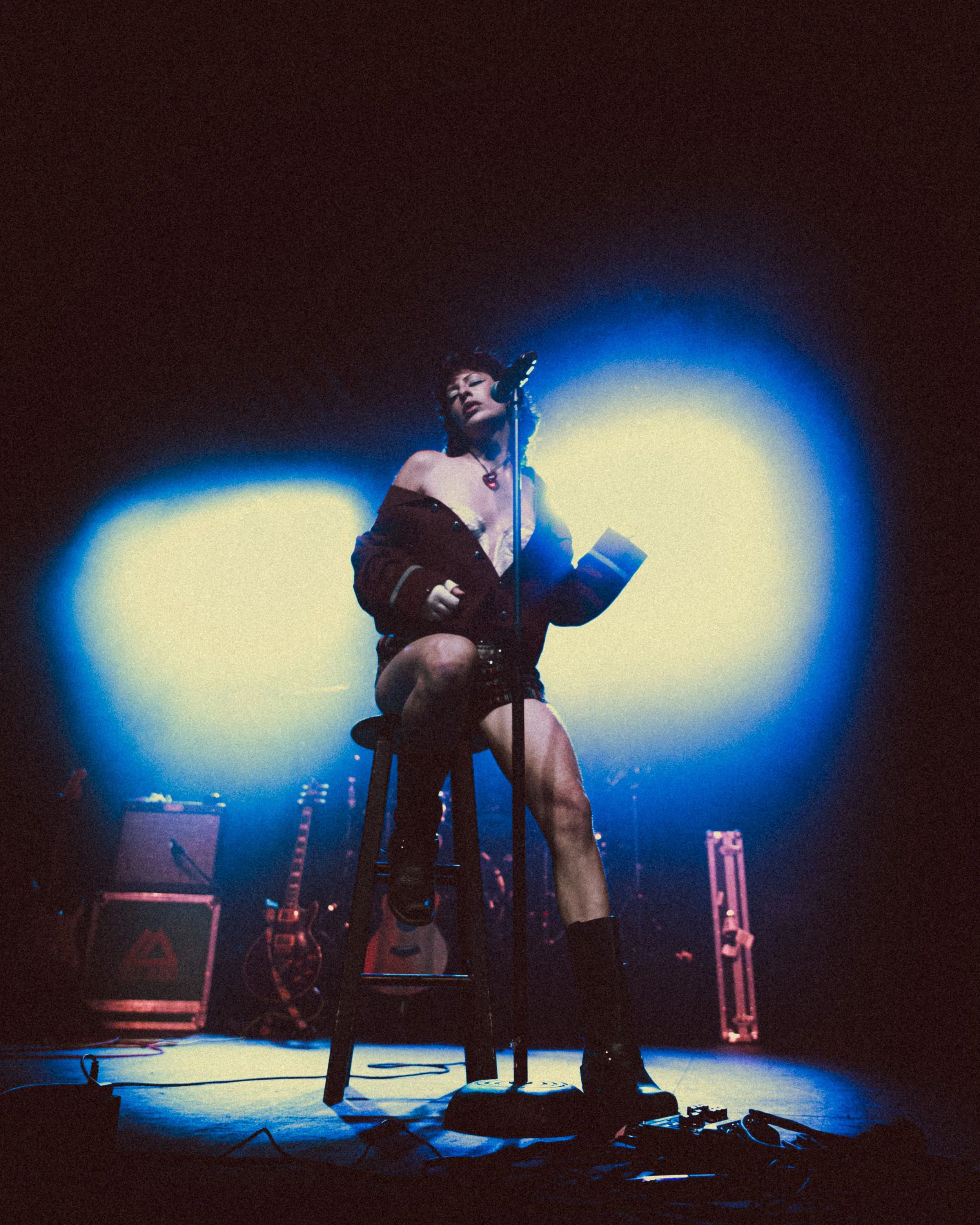 A woman performing on stage under blue and yellow spotlights, sitting on a stool with a microphone, holding a book or paper.