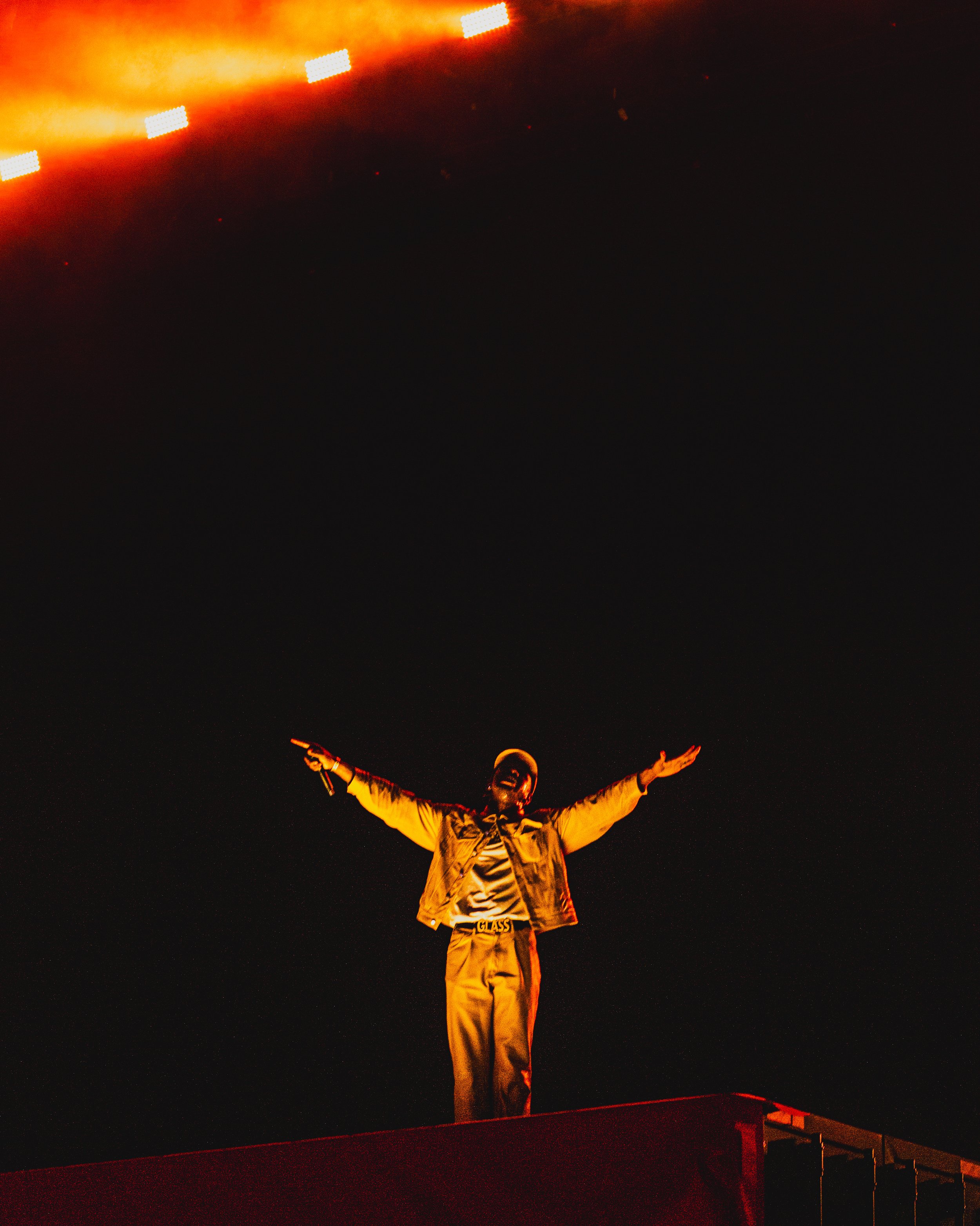 Performer standing on stage with arms outstretched at night, illuminated by orange stage lights.