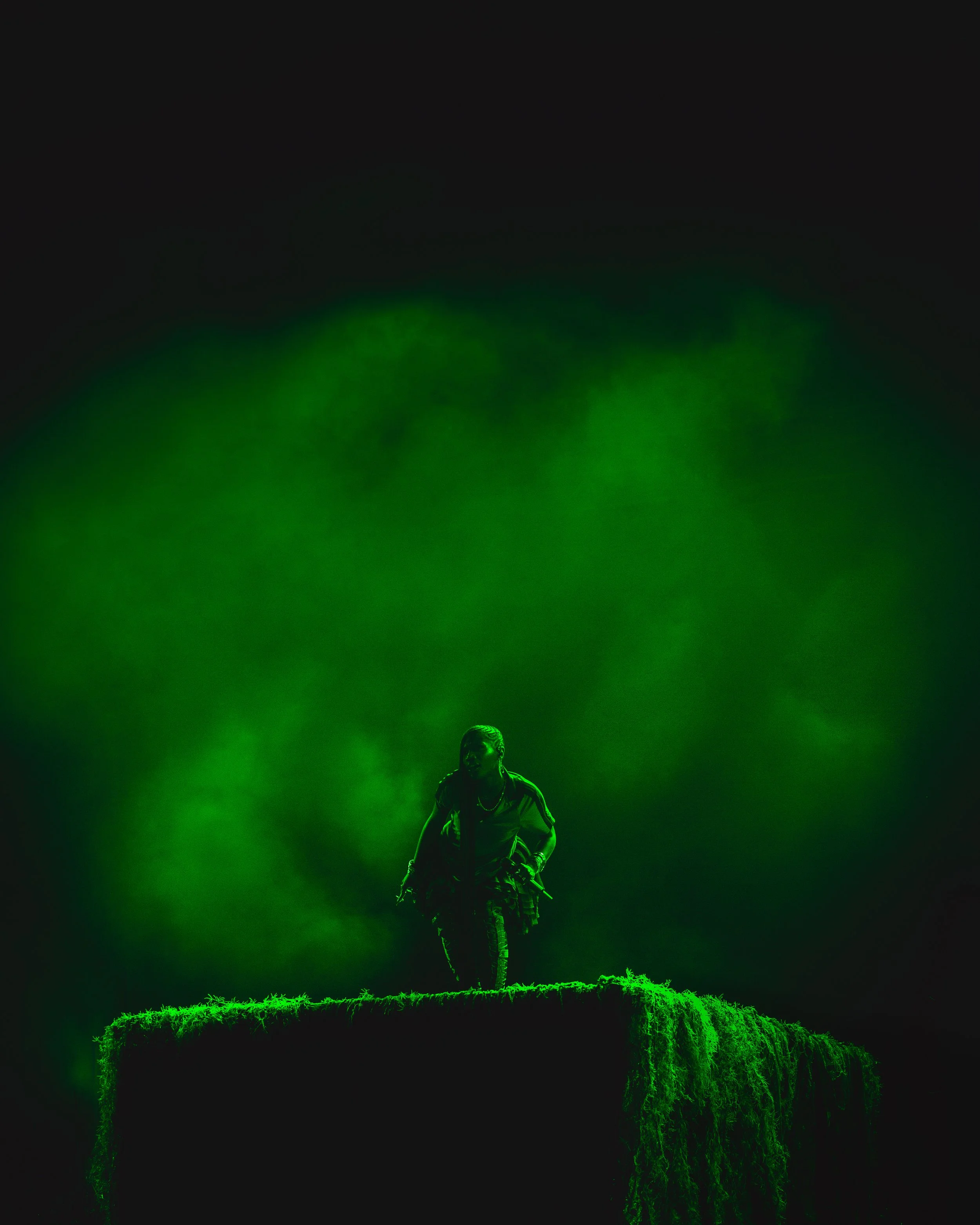 Person standing on a grassy platform at night, illuminated by green lights, with dark sky and green cloud-like smoke in the background.