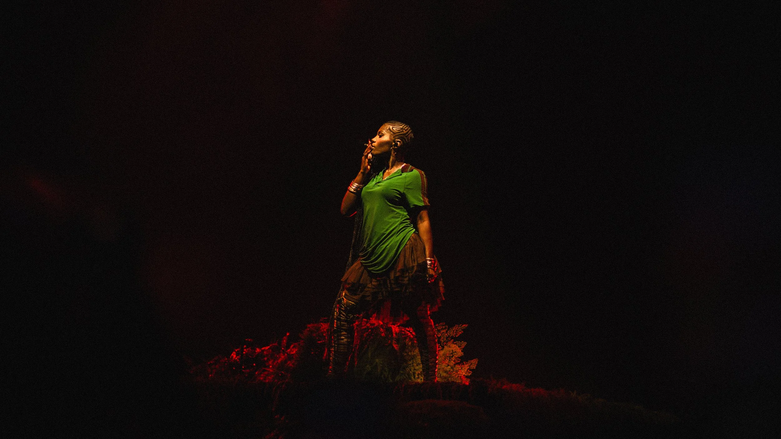 A woman standing on a small hill at night, illuminated by a soft light, wearing a green top, a skirt or dress, and several bracelets, with her hand touching her face.