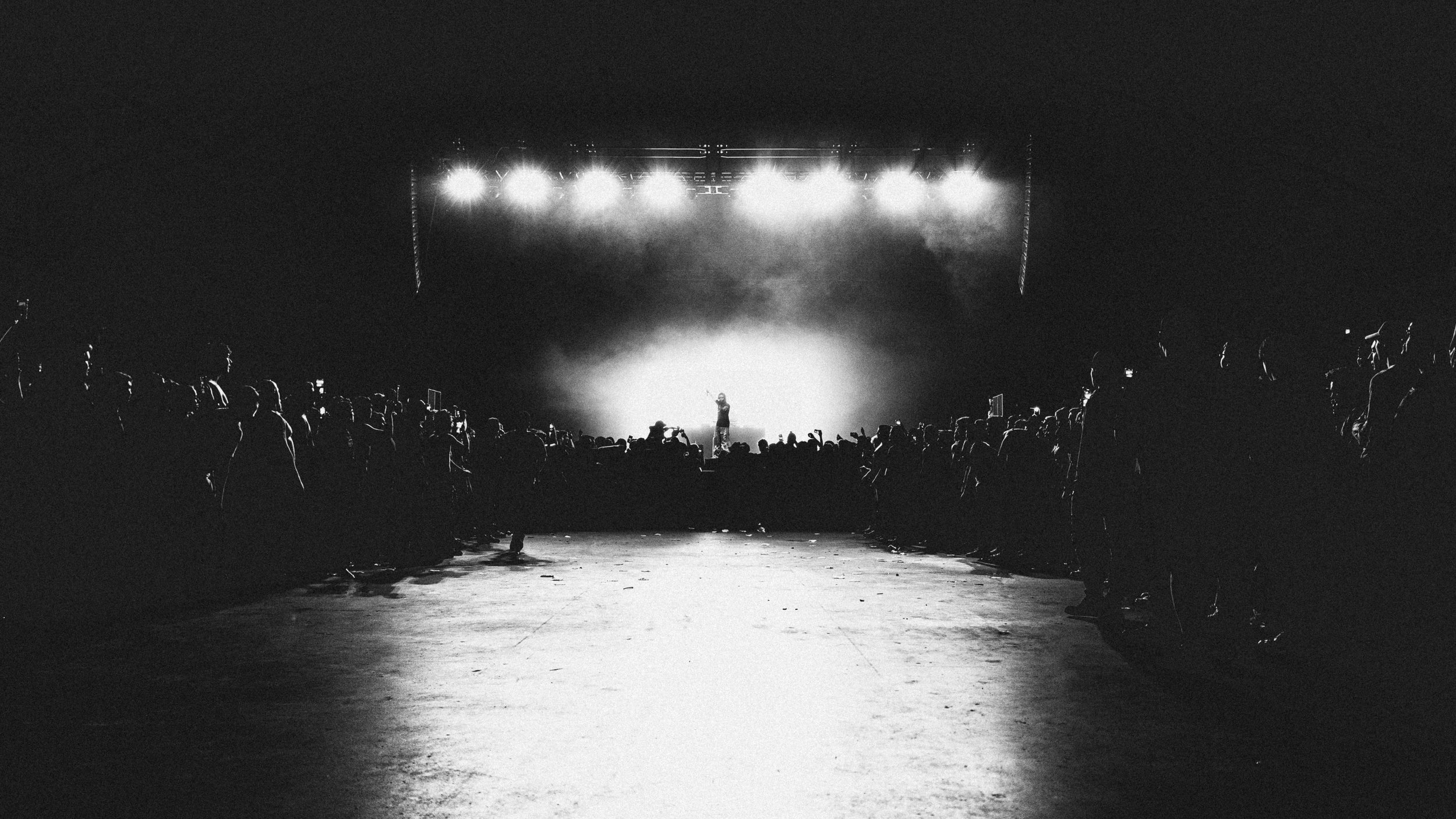 A black and white photo of a concert with an audience facing a brightly lit stage where a performer is standing.