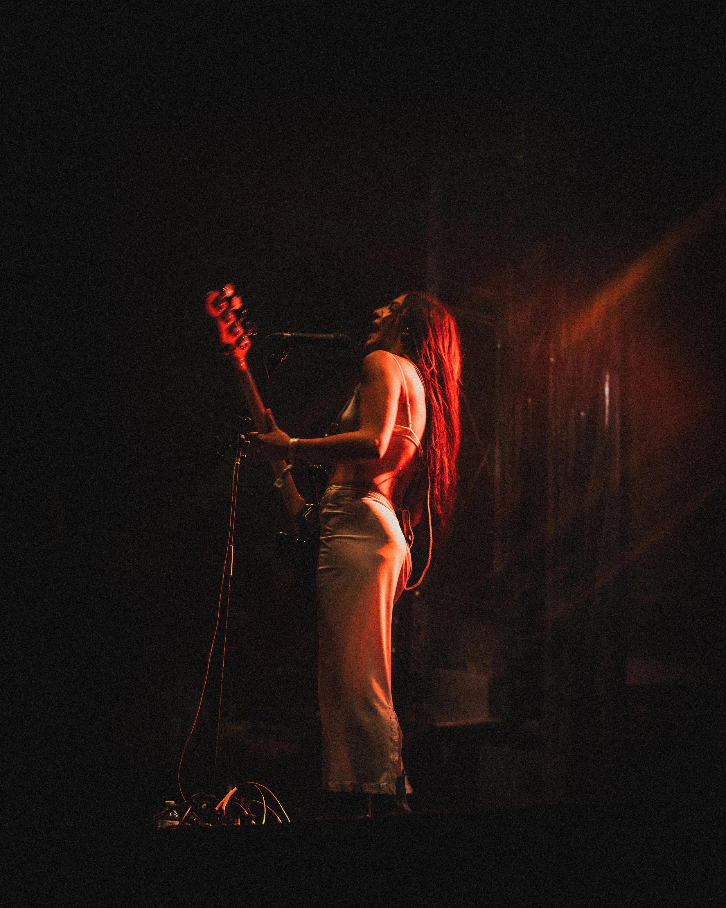 A woman with long hair performing on stage with a bass guitar, singing into a microphone, illuminated by warm stage lights in a dark setting.
