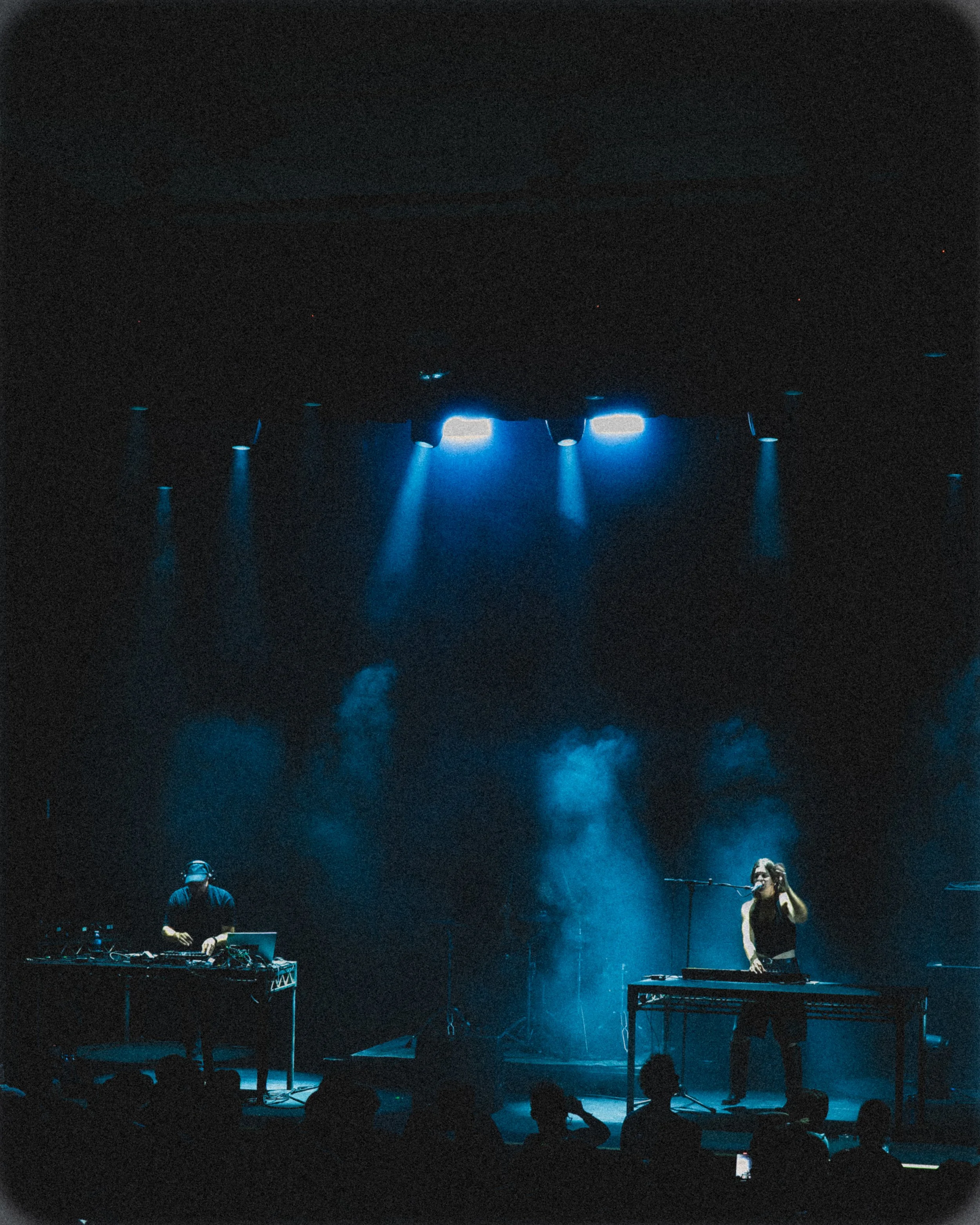 Musicians performing on stage under blue lighting, one using electronic equipment and the other singing into a microphone, with audience silhouettes visible in the foreground.