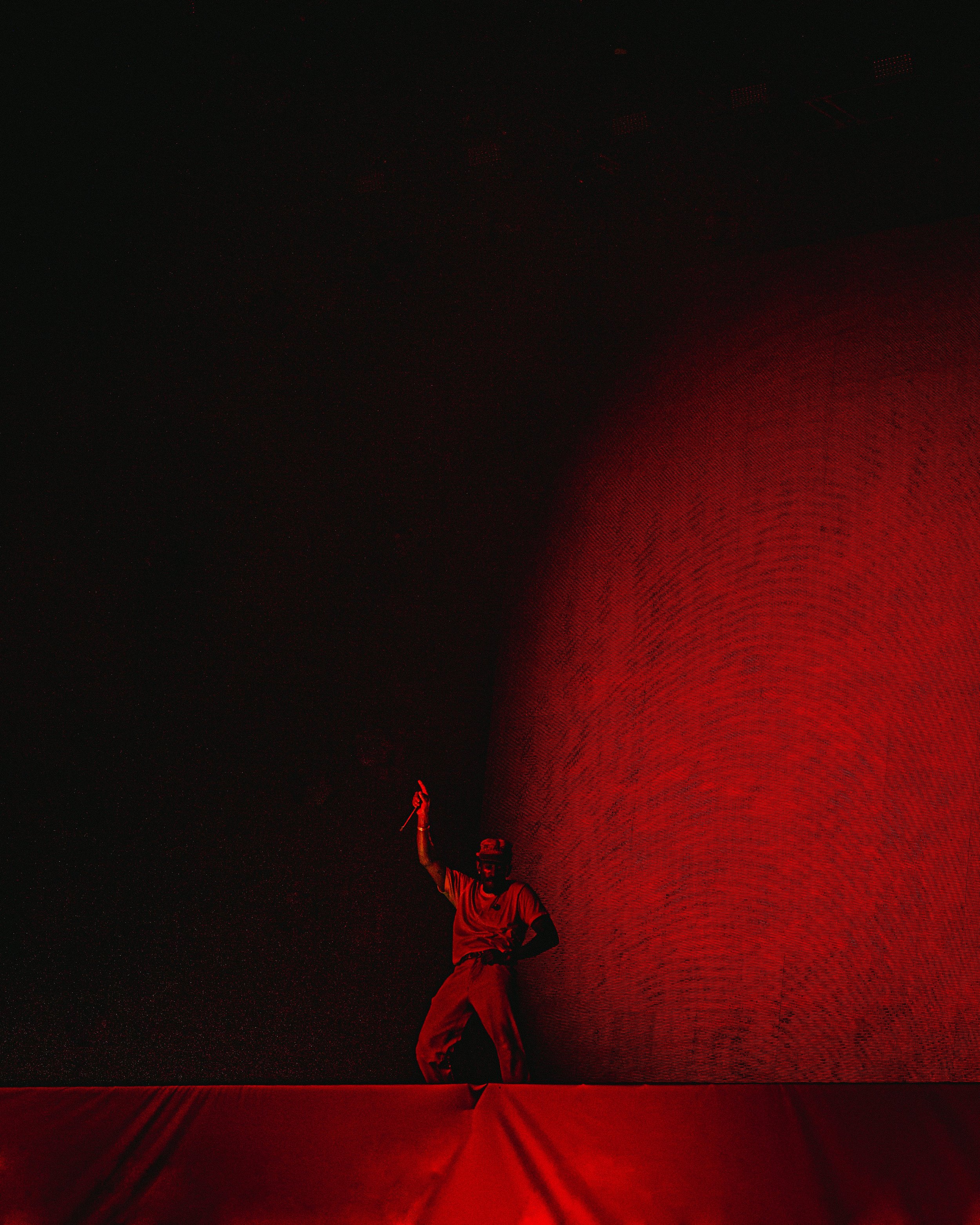 A person dressed in red holding a knife raised in the air, standing in front of a large curved red wall with dark surroundings.