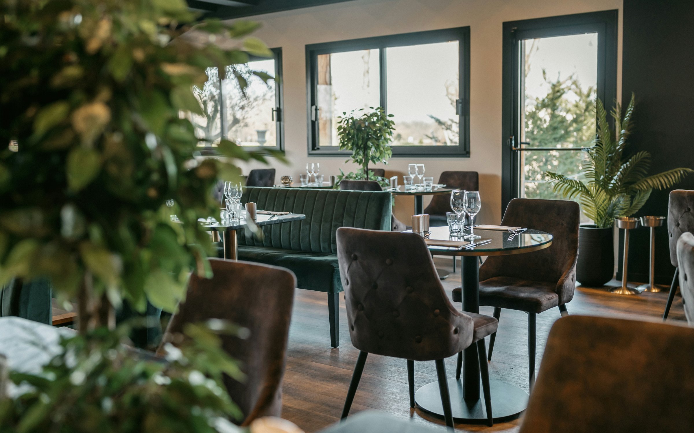 Green upholstered banquette and grey dining chairs around tables beside large restaurant windows.