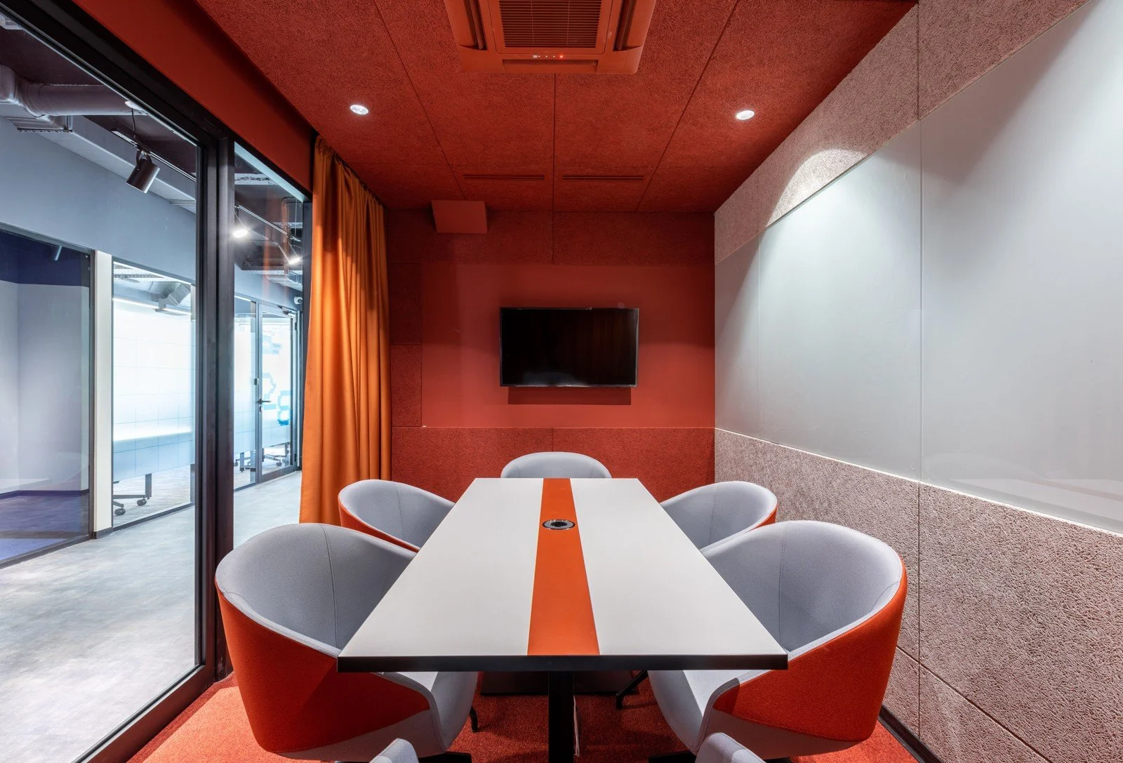 An enclosed meeting room with a glass wall, orange curtain, five chairs, wall screen and ceiling air-conditioning unit.
