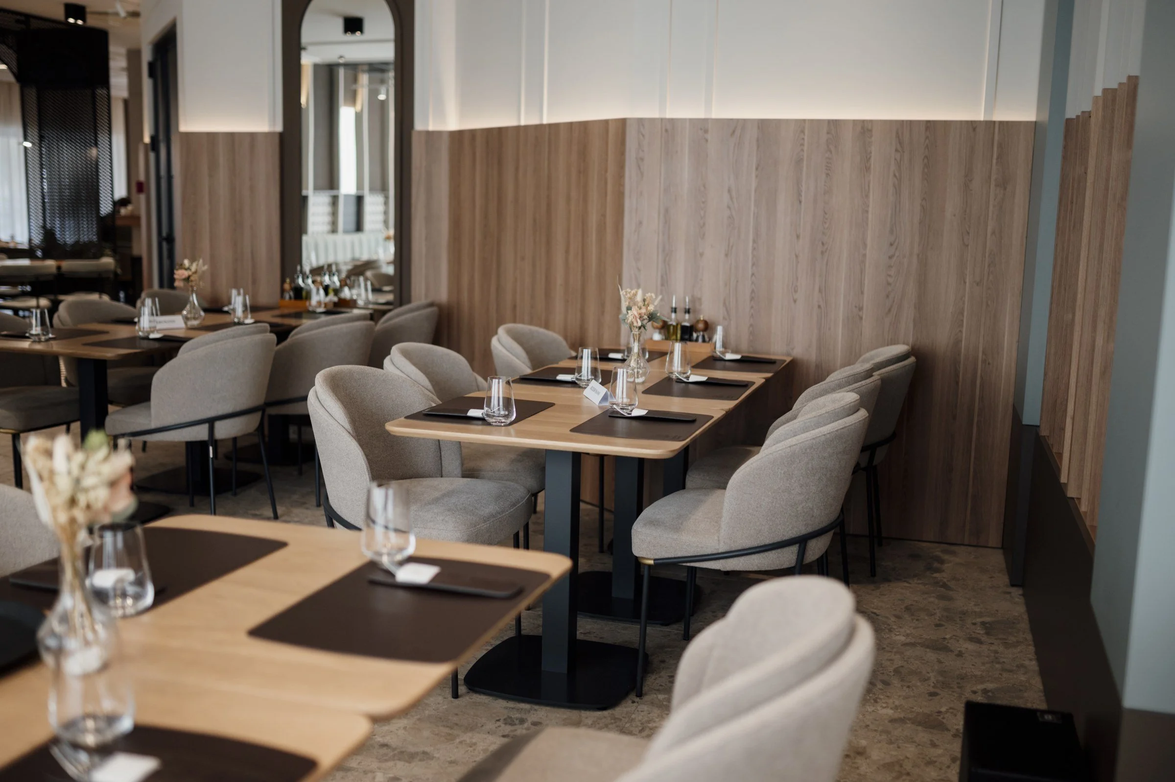 Upholstered dining chairs and closely arranged tables beside timber wall panels and tall mirrors in a restaurant.
