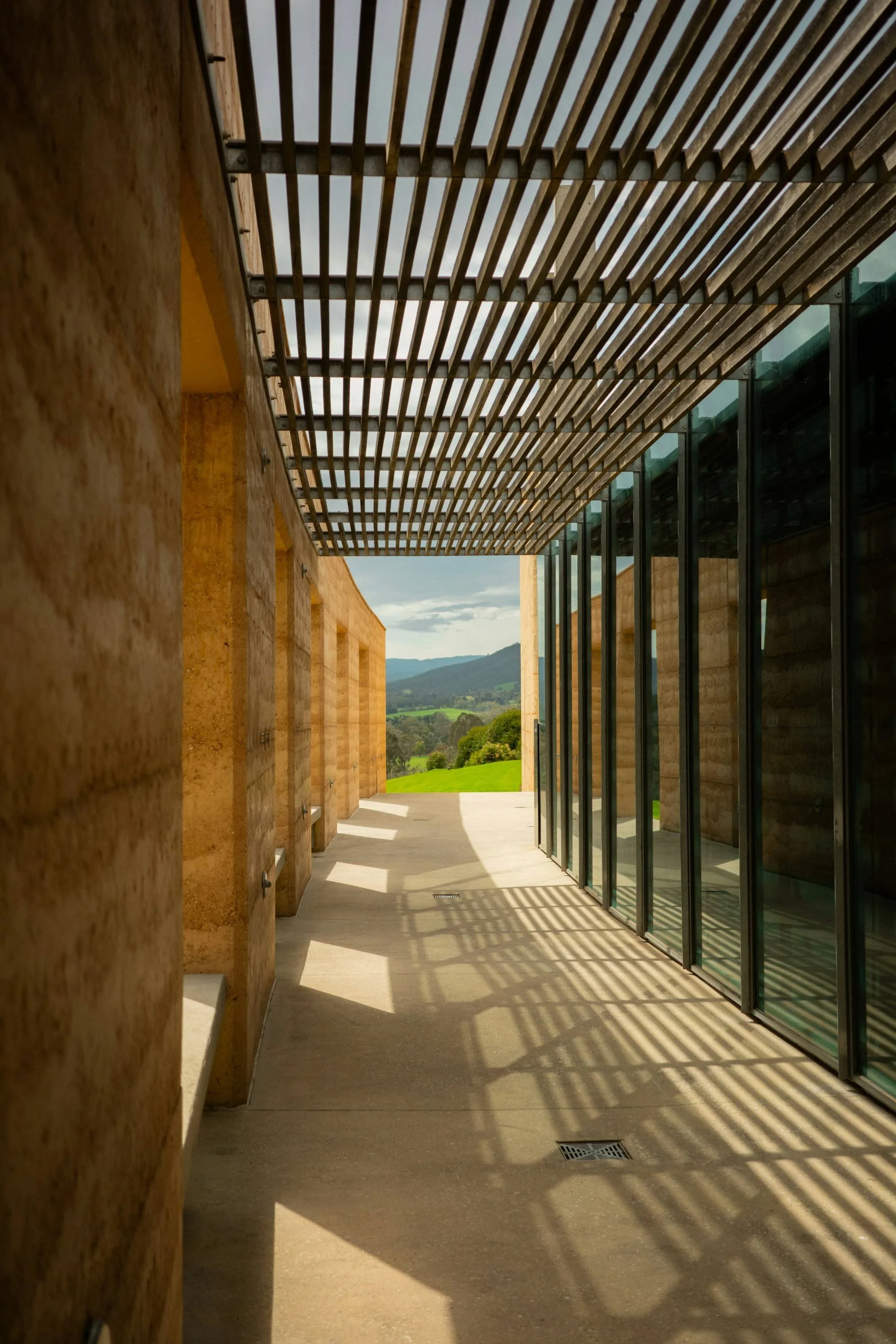Timber slats cast striped shadows along a walkway between warm textured walls and full-height glazing.