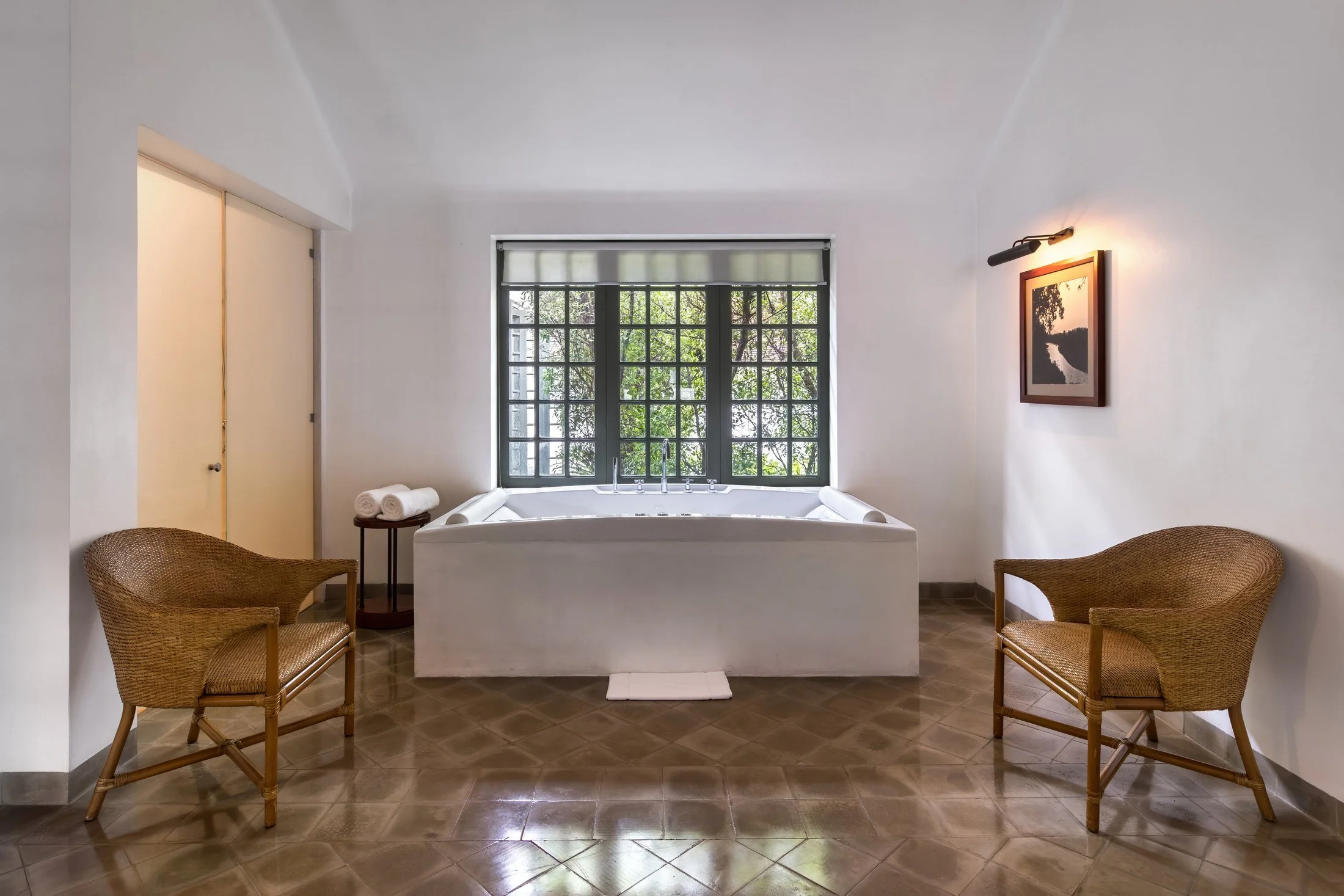 A spa bath beneath a window with a roller blind, with a wicker armchair on either side at Amantaka in Laos.
