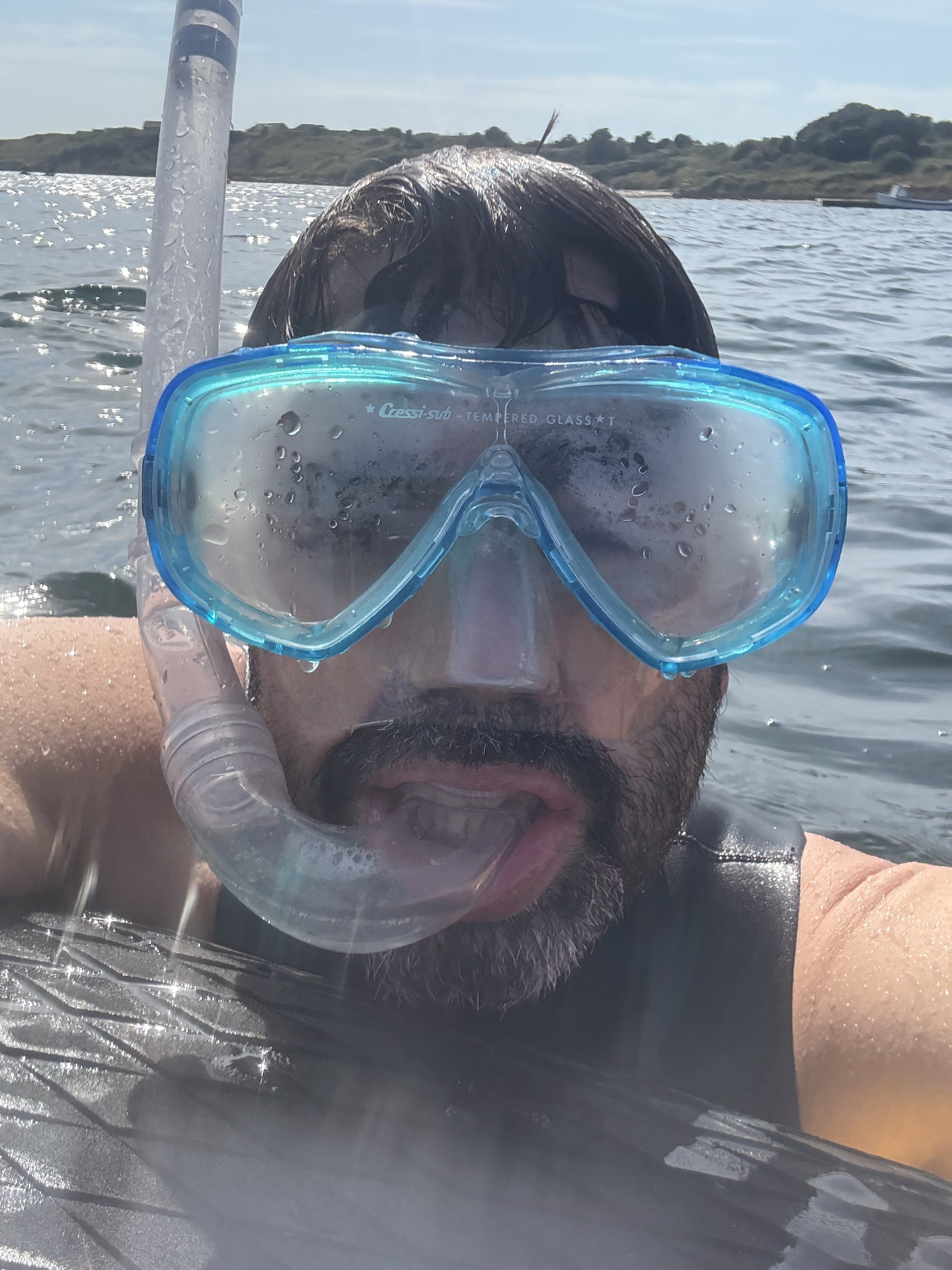 Close-up of a man in swimming gear wearing a snorkel mask, sticking out his tongue, in a body of water with a landscape in the background.