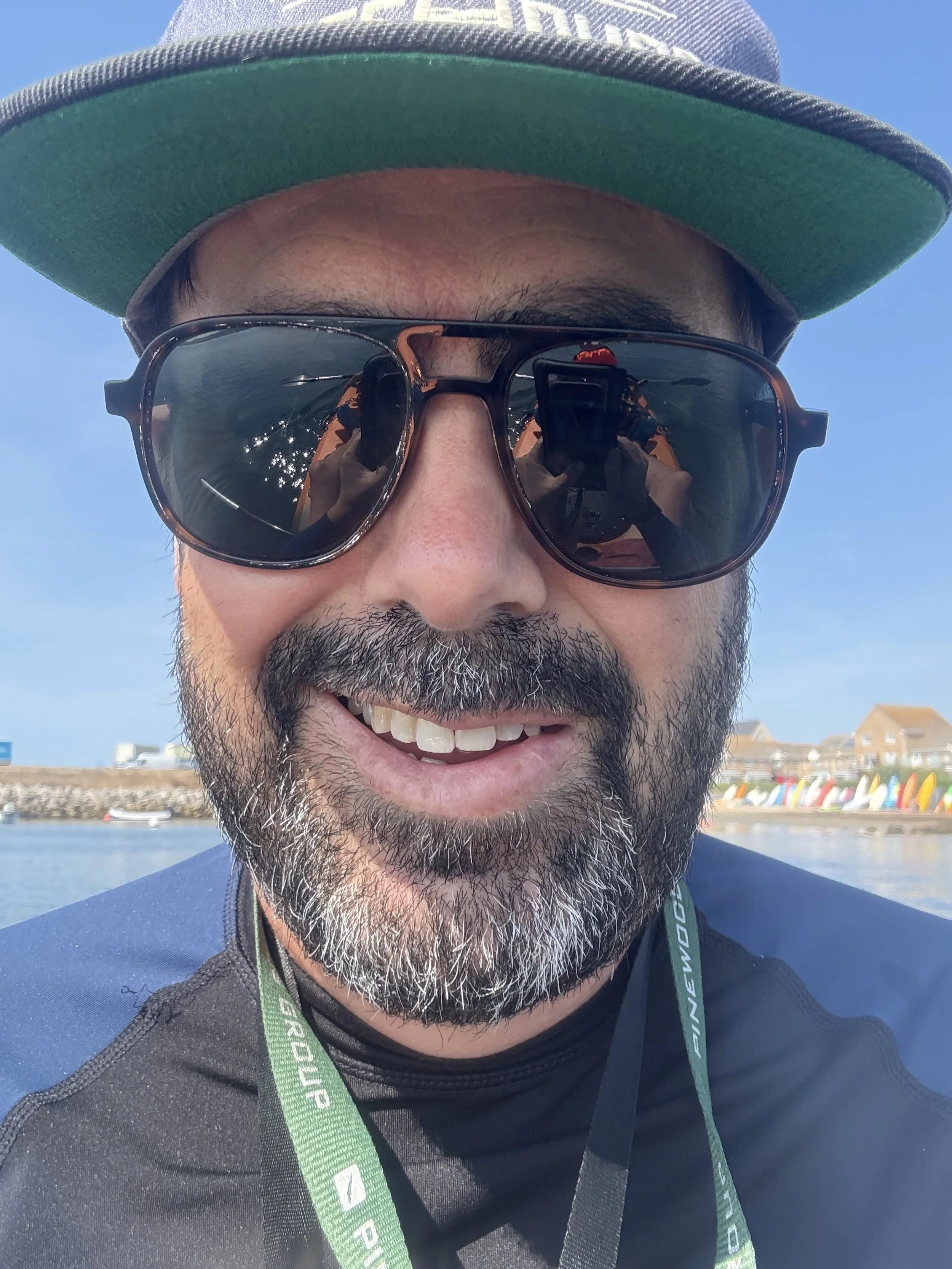 A man with a beard and sunglasses smiling at the camera, wearing a hat and a black shirt, with a body of water and colorful boats in the background on a sunny day.