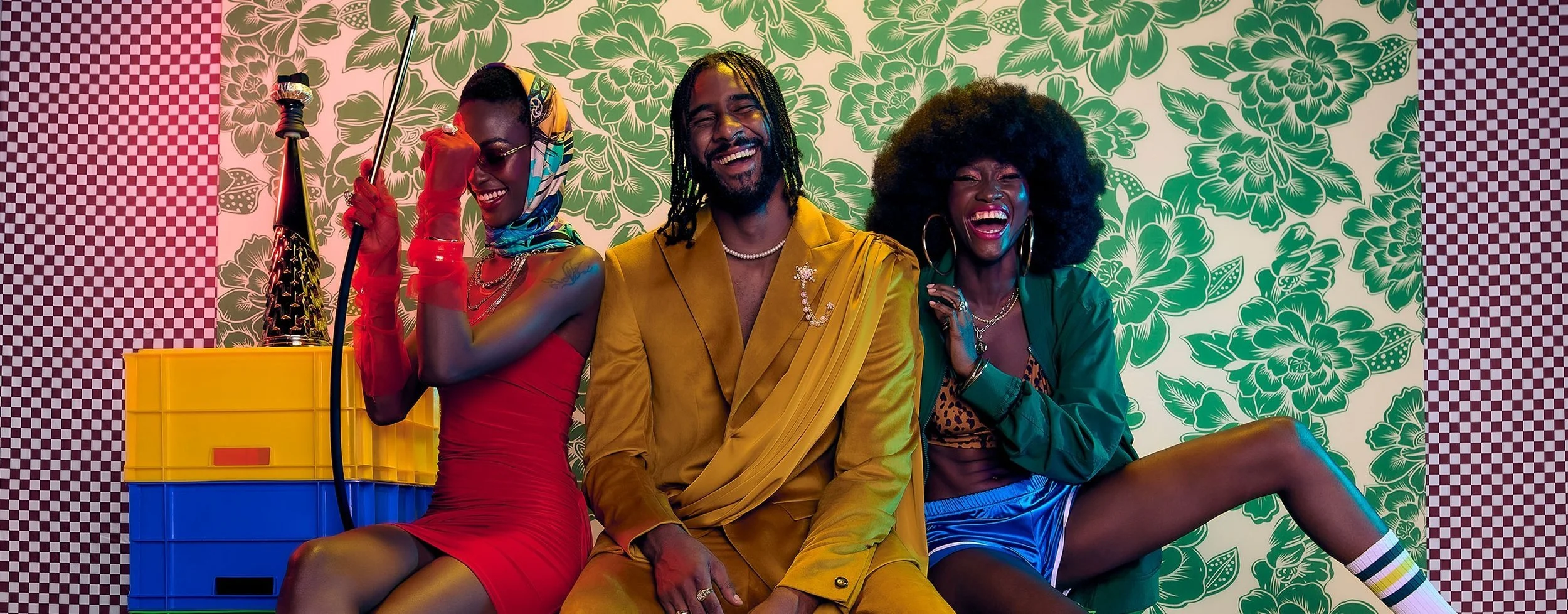 Three diverse people sitting together and laughing against a vibrant floral and checkered background, with colorful clothing and accessories.