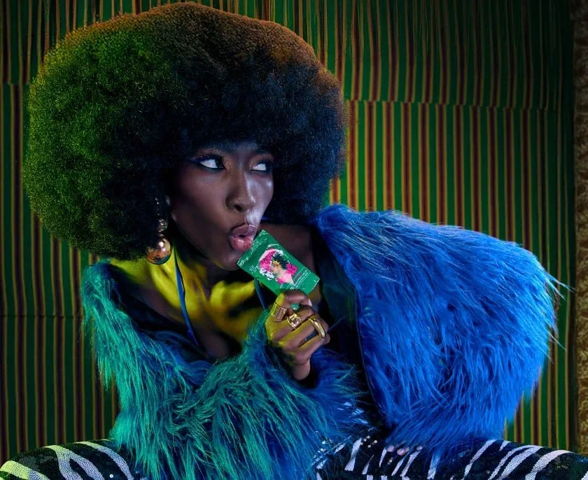 A woman with an afro hairstyle holding a packet of shisha tobacco near her lips, dressed in a vibrant blue fur coat and colorful striped pants, with a striped green and red background.
