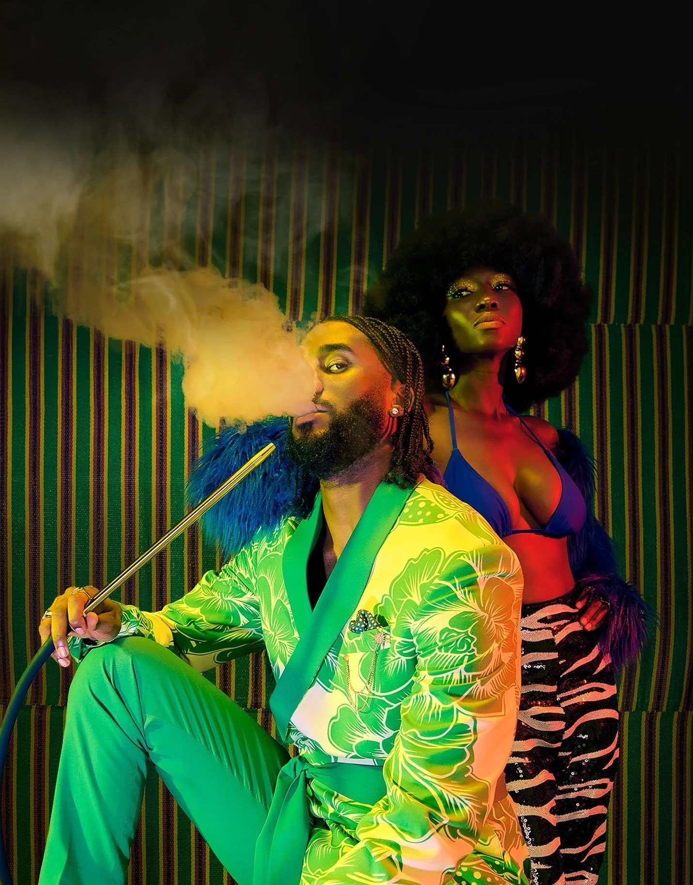 A man with a beard, wearing a vibrant green floral maki, sitting and smoking from a hookah with yellow smoke, and a woman with large afro hair, wearing a blue top and zebra-print pants, standing behind him. Both are in a colorful, striped background.