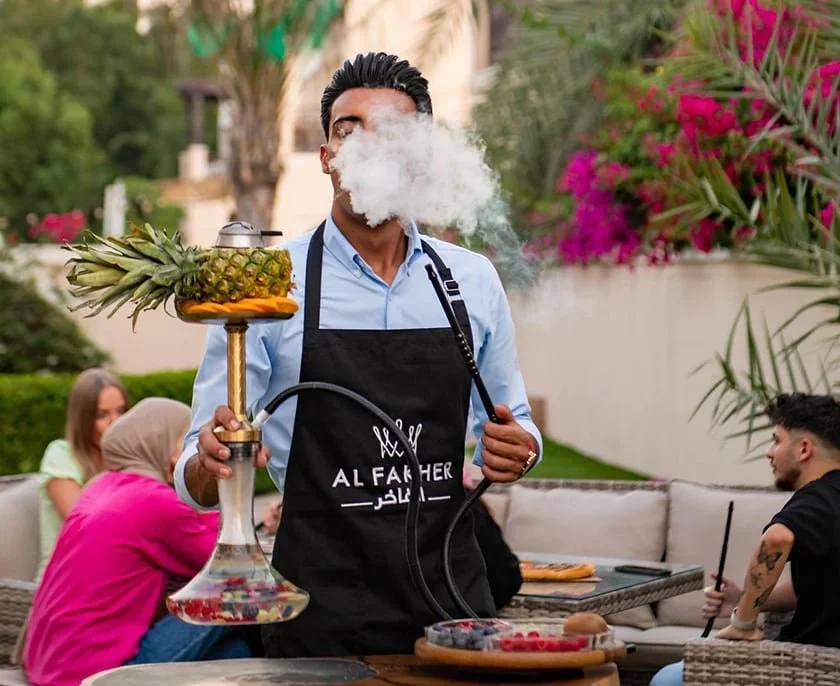 A man in a black apron blowing out smoke from a hookah at an outdoor gathering with people in the background.
