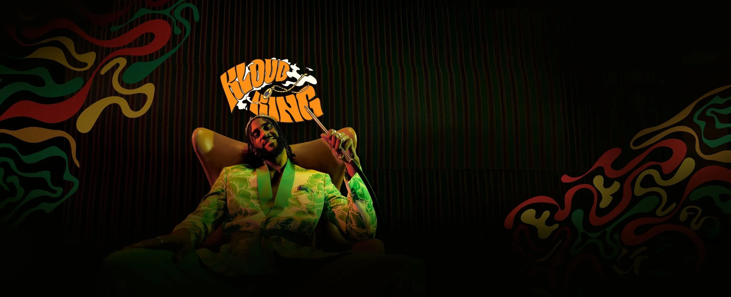 A stylish African man with braided hair, wearing a patterned suit, sitting in a relaxed pose in a dimly lit room with colorful abstract wall art. He holds a sign that says 'KLOUD KING' and is smiling.